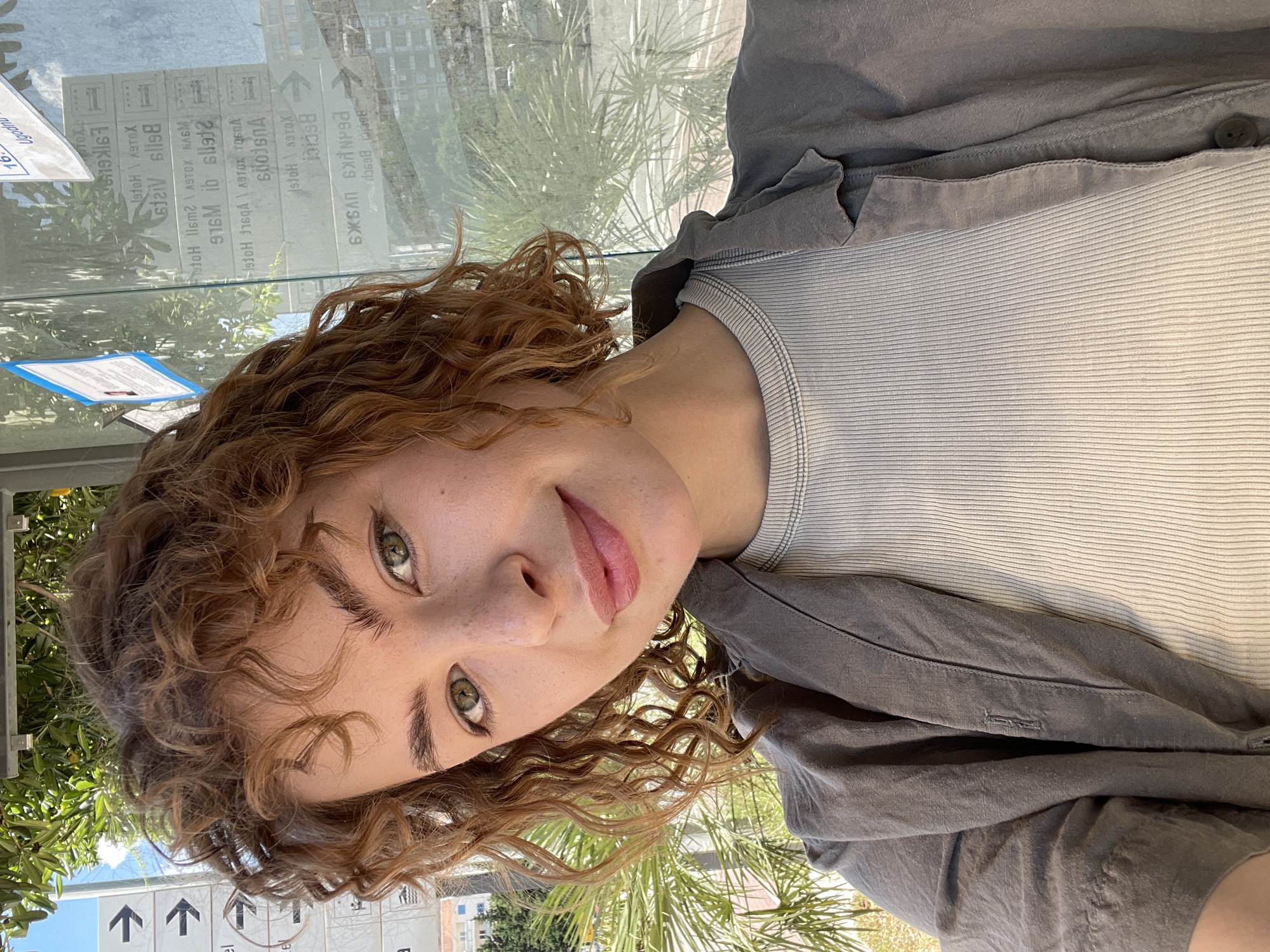 A young woman with curly red hair, green eyes, and light skin is the primary subject of the image. She is wearing a light gray, vertically-ribbed tank top, over which is a darker gray, collared shirt.
The background is composed of an outdoor scene with palm trees and buildings. There is also a bus stop with signs indicating destinations, including "Becici." Based on the destination sign, this image appears to be in the city of Rafailovići, Montenegro.