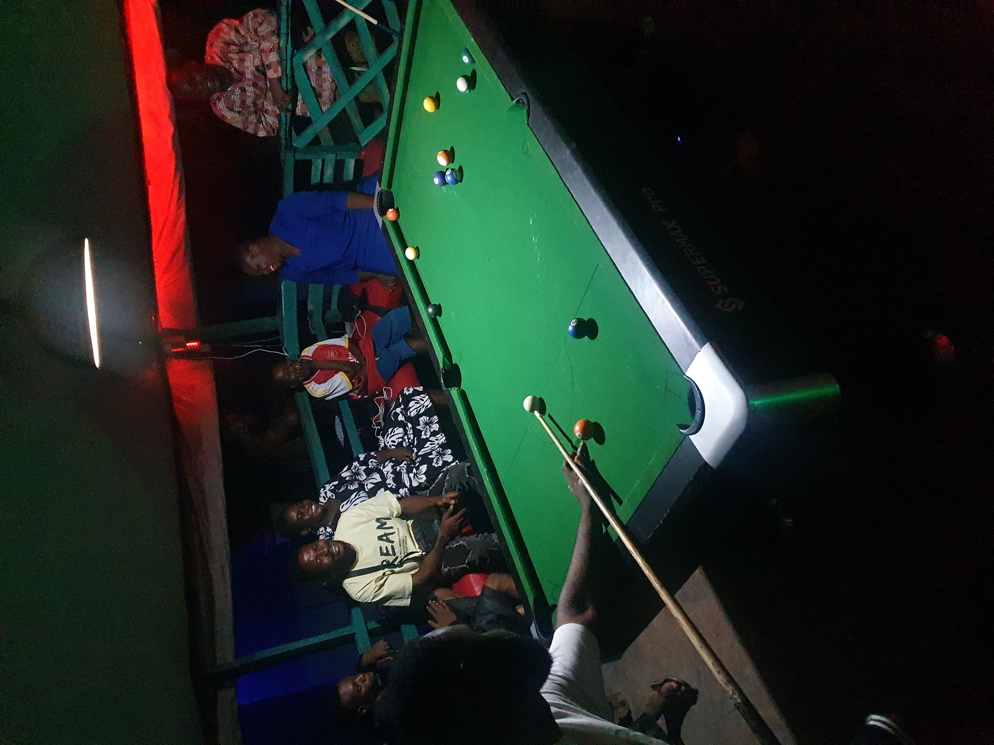 The image depicts a nighttime scene centered around a green pool table in Lokoja, Nigeria. Several people are positioned around the table, some sitting and others standing, appearing to be observing or participating in a game of pool. The pool table has multiple billiard balls scattered across its surface. One person is actively holding a pool cue, seemingly preparing to take a shot. The environment includes a structure with what appears to be wooden supports and a mix of artificial lighting, including a strip light and colored lights that illuminate the area. The lighting is somewhat dim and uneven. A person is lying prone on the adjacent structures, along with other onlookers.