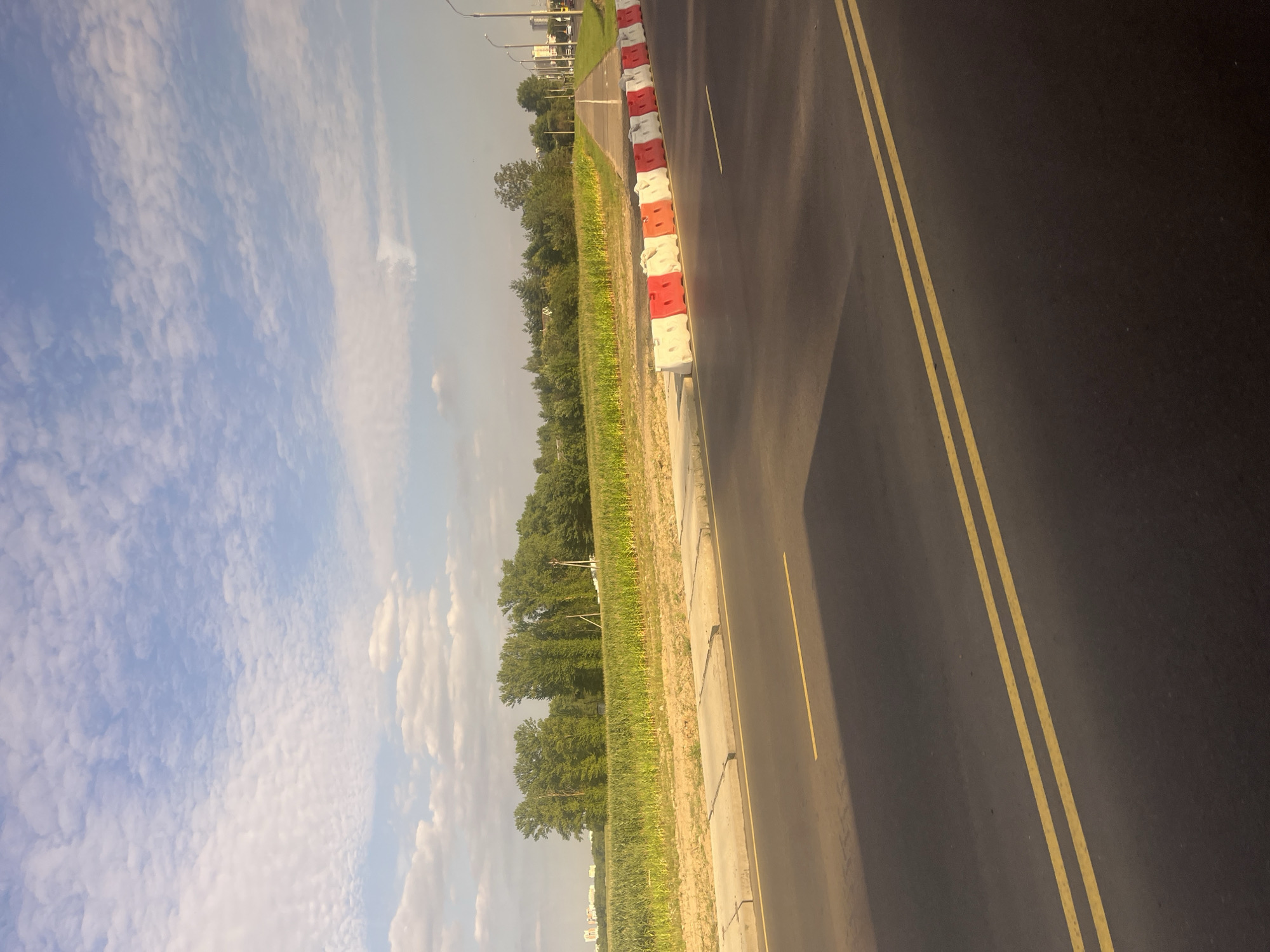 The image captures a view of a paved road with double yellow lines, leading into the distance. On the right side of the road, a row of white and red plastic barriers demarcates a construction or closed-off area. Beyond these barriers, a pathway or sidewalk is visible, followed by a field of green crops, possibly corn, bordered by a line of trees. In the far distance, on the left side of the frame, a cityscape with buildings can be observed. The sky above is a pale blue with scattered white clouds, suggesting daytime. The scene appears to be an urban or suburban fringe area. The location is identified as Minsk, Belarus, likely along a road undergoing development or with restricted access. No people are present in the image, and no actions or interactions are depicted.