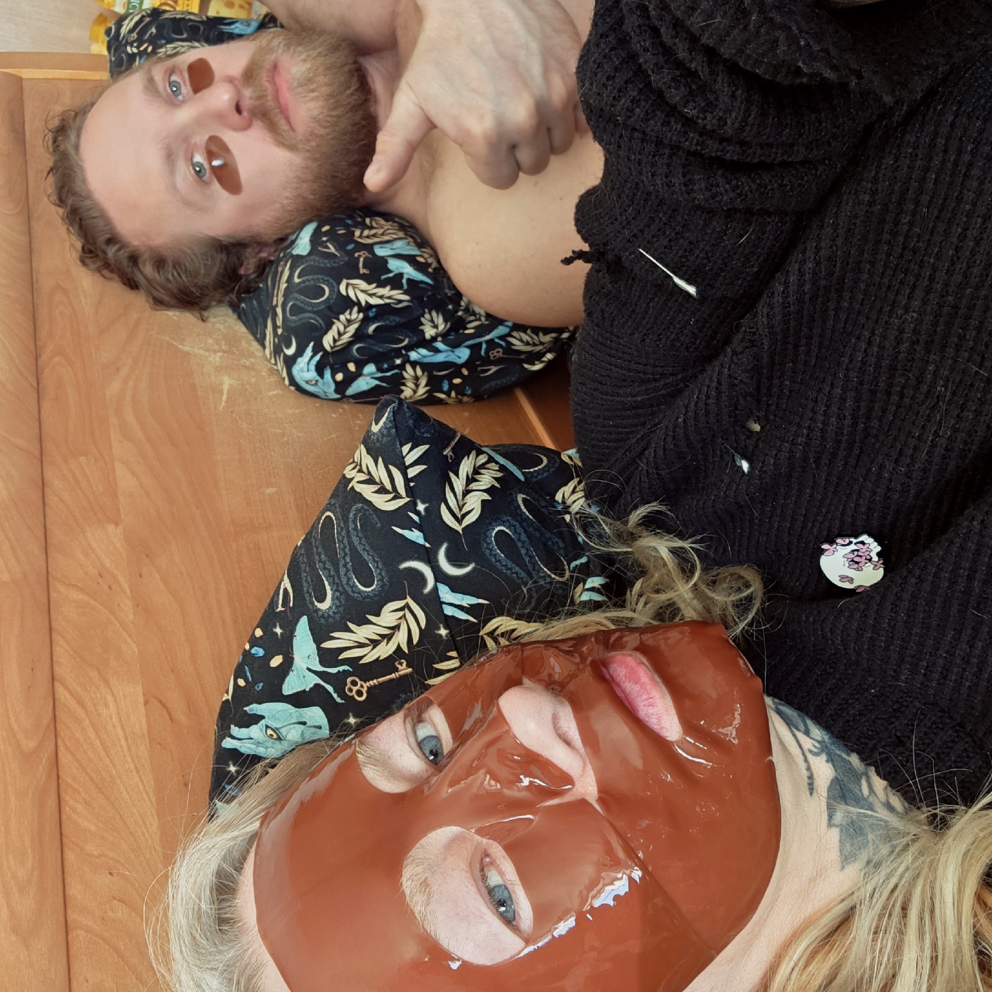 Two individuals are lying on a wooden surface, possibly a floor. One person has a beard and is wearing under-eye patches and patterned clothing with images of keys, creatures, and moons. The second person has a red face mask on and is wearing a dark sweater with a pin showing a white face with cherry blossoms. The sweater has a loose thread. The person with the beard is pointing with his finger. The individuals appear to be posing for the photo. The location in the city of Saint Petersburg, Russia cannot be determined from the image.