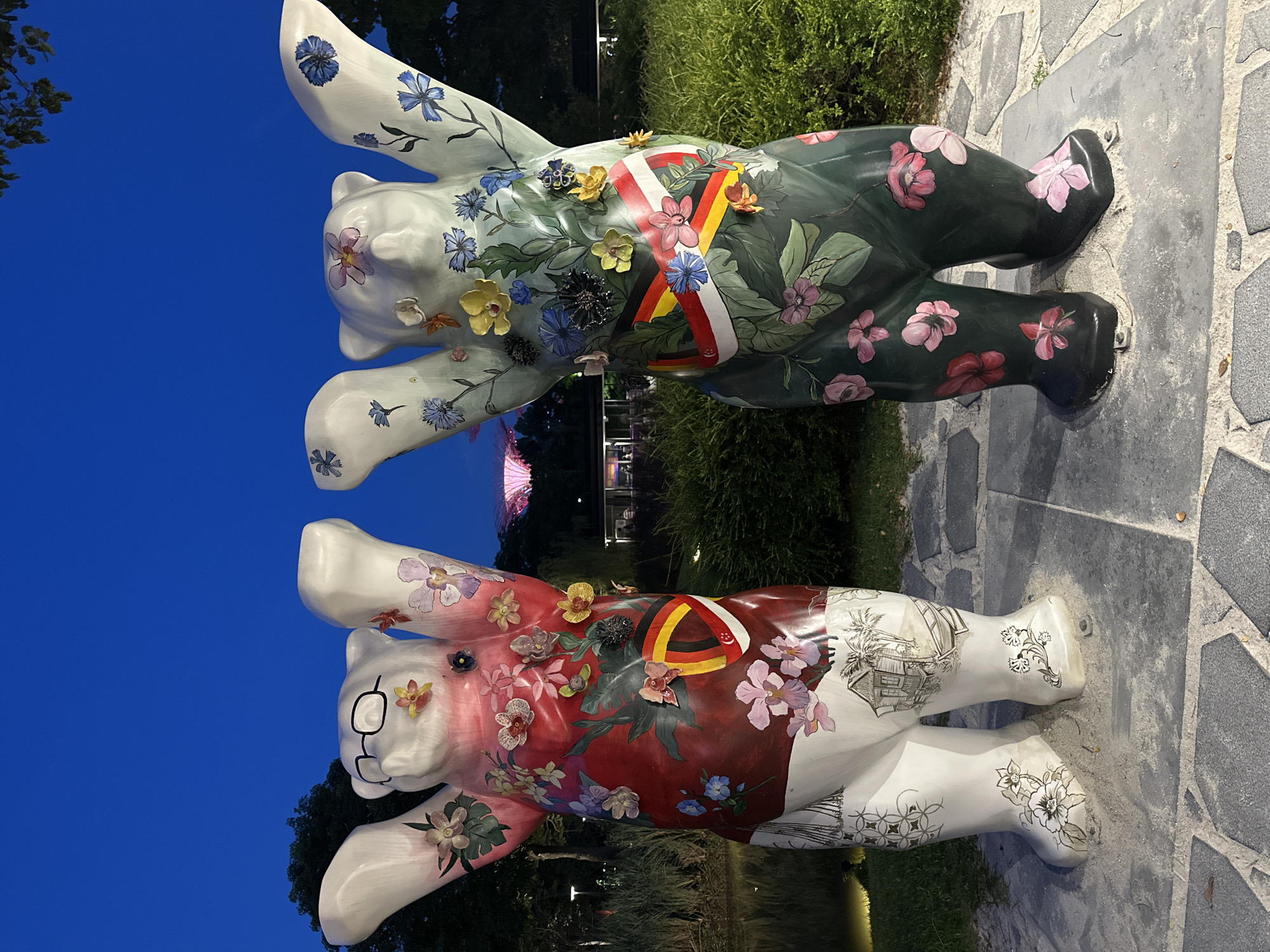 This image depicts two large bear sculptures in the Gardens by the Bay in Singapore. The sculptures are positioned in the foreground, standing on a paved walkway, with their arms raised. The sculpture on the left is white with red accents and adorned with various flowers, glasses, and a sketch of a house with a boat. The sculpture on the right is dark green and also decorated with flowers. Both sculptures have bands around their torsos, one featuring the colors of the German flag and the other featuring the Singaporean flag. In the background, the iconic Supertrees of Gardens by the Bay are visible, illuminated against the twilight sky. The surrounding environment consists of lush greenery and a body of water.