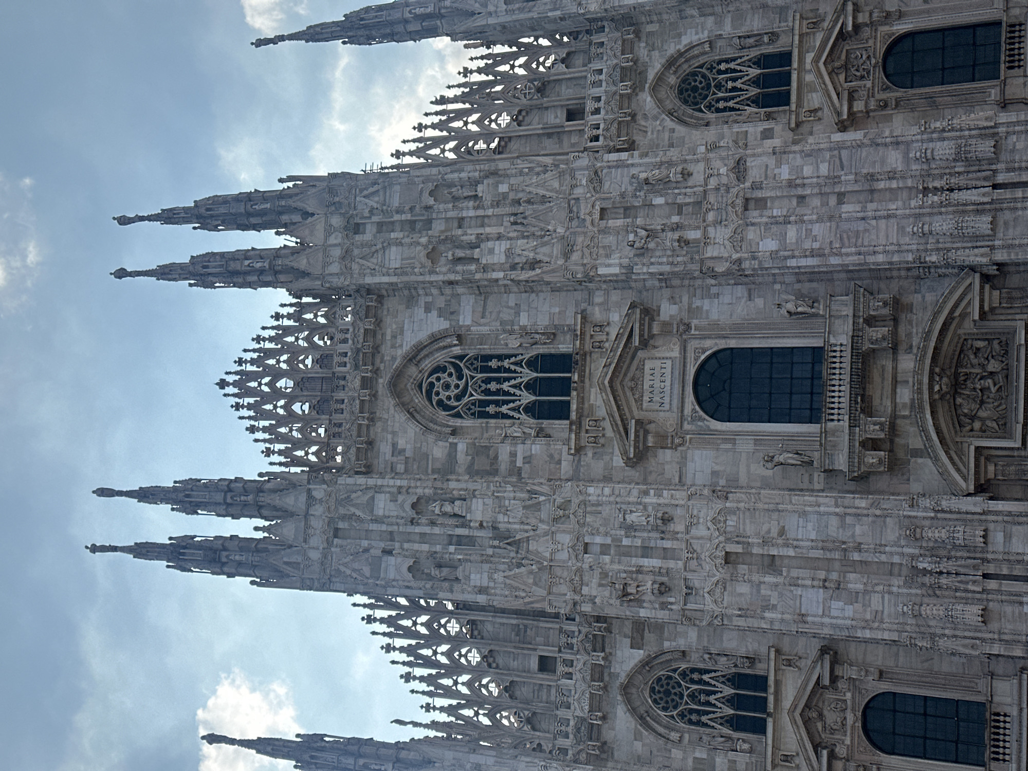 The image shows a close-up, frontal view of the facade of the Duomo di Milano (Milan Cathedral) in Milan, Italy. The cathedral is constructed of white marble and features intricate Gothic architecture with numerous spires, statues, and ornate carvings. The sky above is cloudy with patches of blue. The image is taken from a low angle, emphasizing the height and grandeur of the building. No people are visible in the frame. The architectural style is characterized by pointed arches, ribbed vaults, and flying buttresses, although the latter are not fully visible in this frontal shot. The facade is divided into several vertical sections by buttresses and adorned with a profusion of sculptures and pinnacles. The lighting suggests it is daytime.