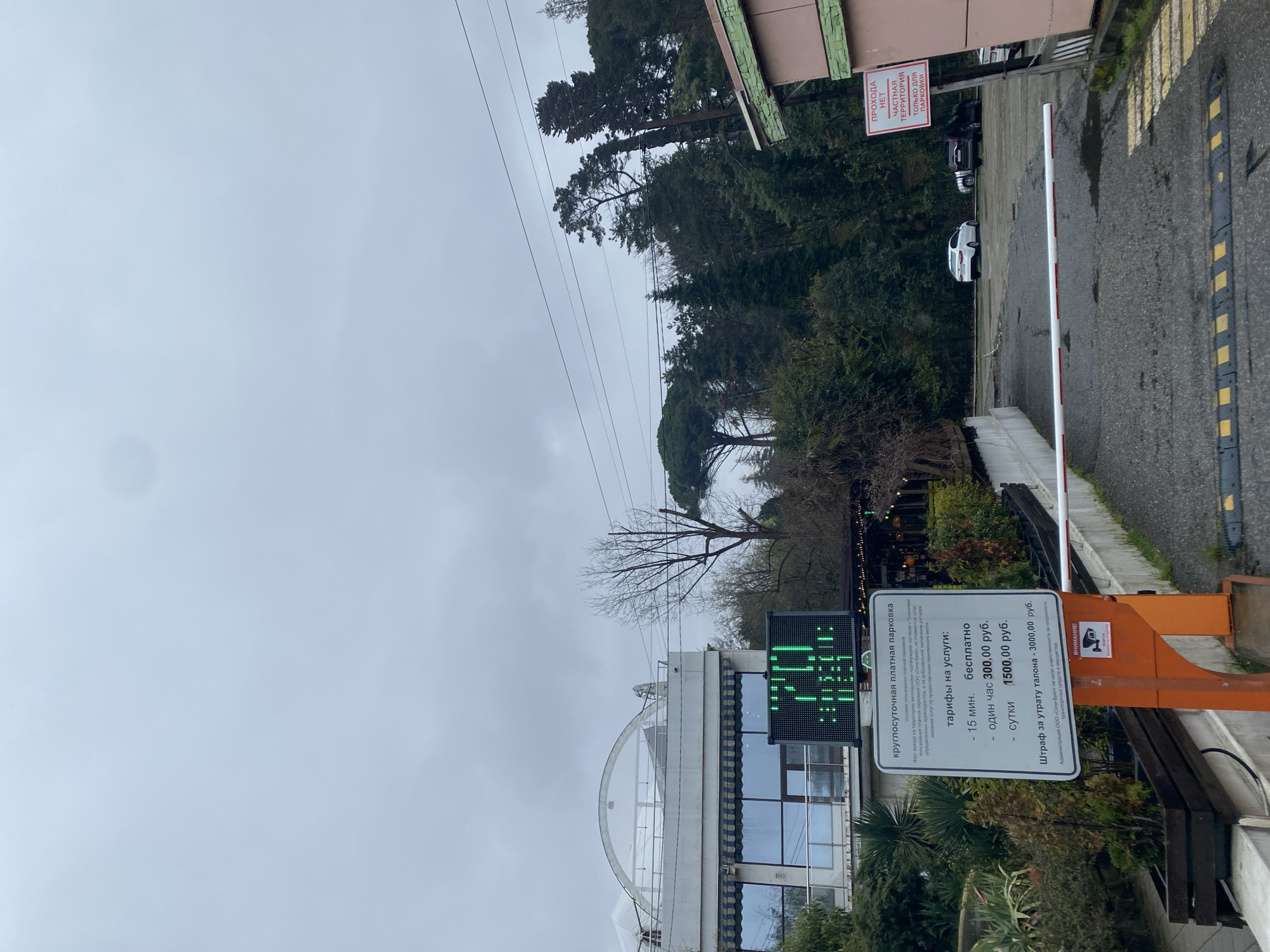 The image depicts an entrance to a paid parking area in Sochi, Russia. The entrance is marked by a white and red striped gate. A sign displays parking rates: 15 minutes are free, one hour costs 300 rubles, and a full day costs 1500 rubles. A fine of 3000 rubles is noted for lost parking tickets. A sign states, "No passage. Private territory for parking only." The area seems to be on a hill, as trees are seen above a building with a white dome. A couple of cars are seen parked beyond the gate. The weather appears overcast.