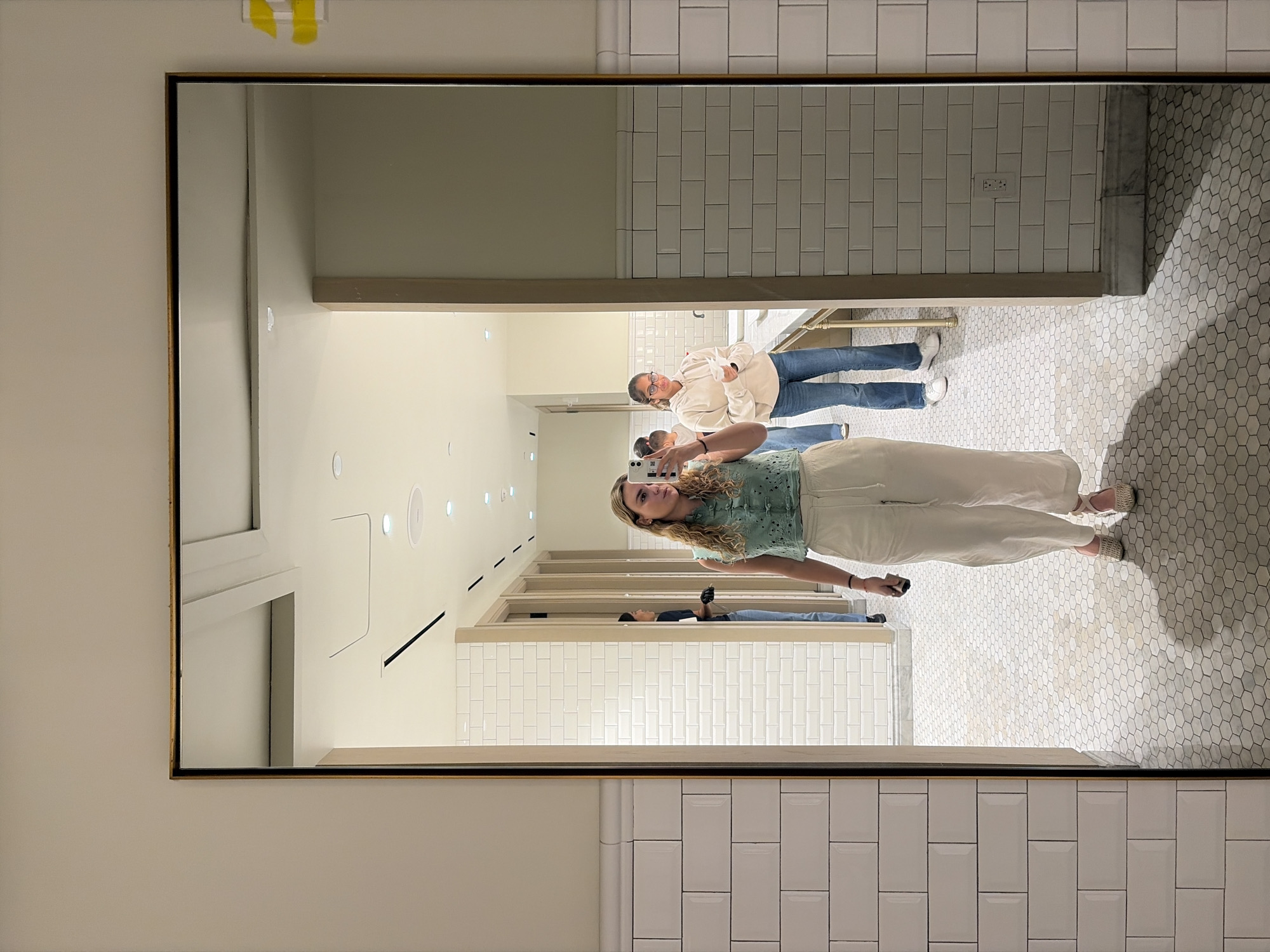 The image depicts a mirror reflection in a bathroom, likely public due to the communal sinks visible in the background. Three individuals are present. The main subject is a woman taking a selfie in the mirror, wearing a green shirt and light-colored pants. Behind her and to the left, another woman wearing glasses and a white sweatshirt is standing at the sink. Further in the background and further left, a third person is also present. The bathroom features white tiled walls and a hexagonal-patterned tiled floor. The setting has a modern and clean aesthetic. The precise location in Miami, United States of America, cannot be conclusively determined from the provided visual information alone.