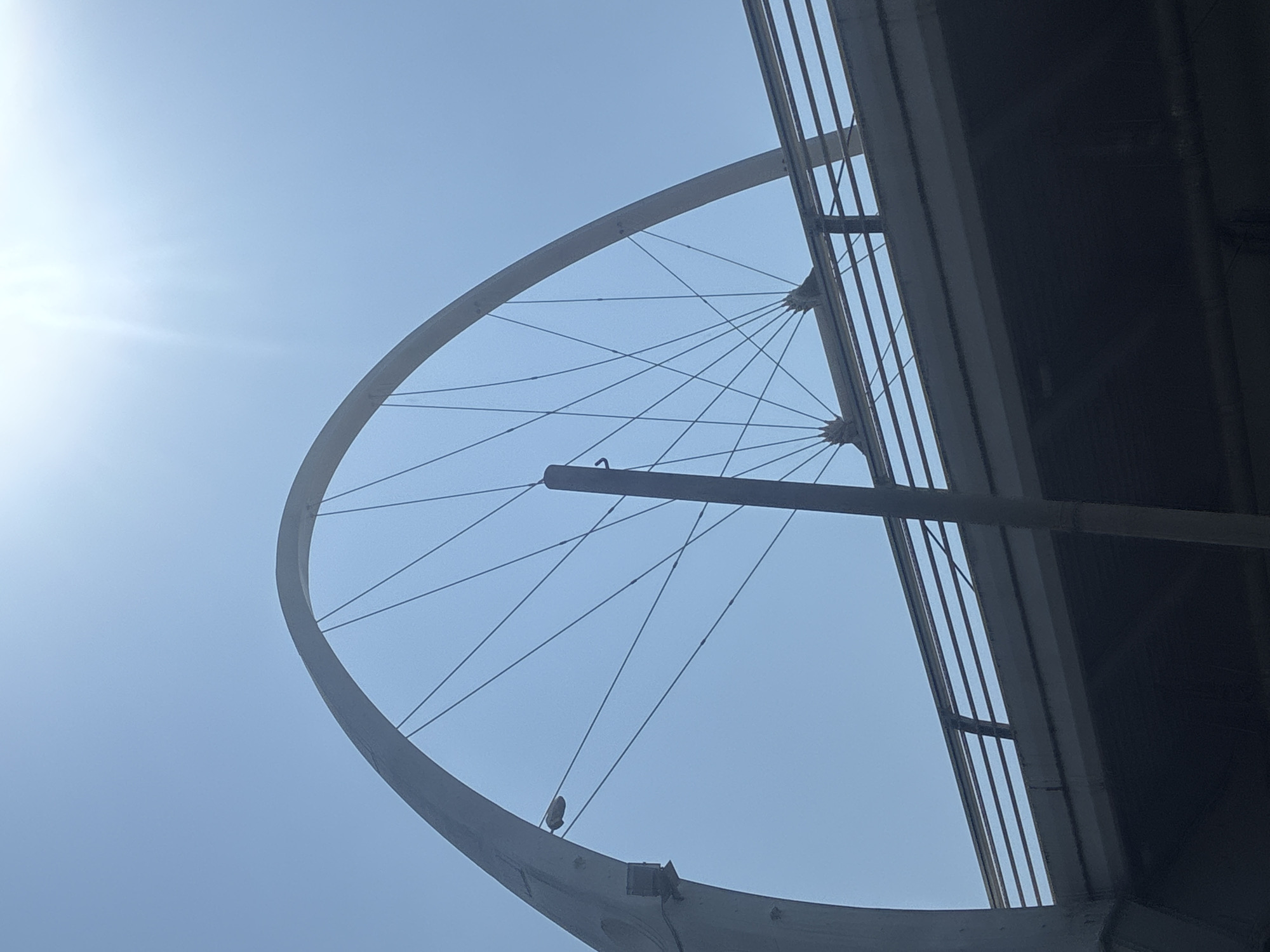 The image depicts a view from below of a pedestrian bridge with an arched, spoke-like structure against a bright, clear sky. The sun is visible in the upper right corner, creating a lens flare effect. The bridge has a metal railing running along its length. The arched structure resembles a large wheel with cables extending from the apex to the rim. Two figures are partially visible standing on the bridge, silhouetted against the sky. The overall scene is brightly lit, suggesting daytime. The location is identified as the Millennium Bridge in Sochi, Russia.