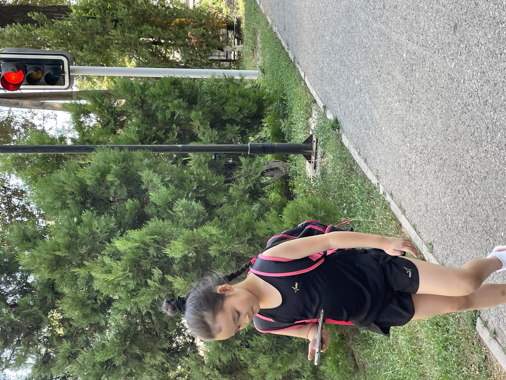 A young girl, appearing to be of Asian descent, walks along a paved pathway bordered by grass and dense green trees. She has dark hair pulled up in a bun with a braid trailing down her back and is wearing a black sleeveless athletic top with pink trim and black athletic shorts. She carries a black backpack with pink accents and holds a silver smartphone in her left hand, looking down at it. To her right, a traffic light with a red signal illuminated is mounted on a white pole, alongside a dark vertical pole. The background consists of lush green foliage from trees and other vegetation, with a hint of a street or fence visible behind them. The scene appears to be an outdoor, urban environment within Almaty, Kazakhstan, likely a park or a tree-lined street.