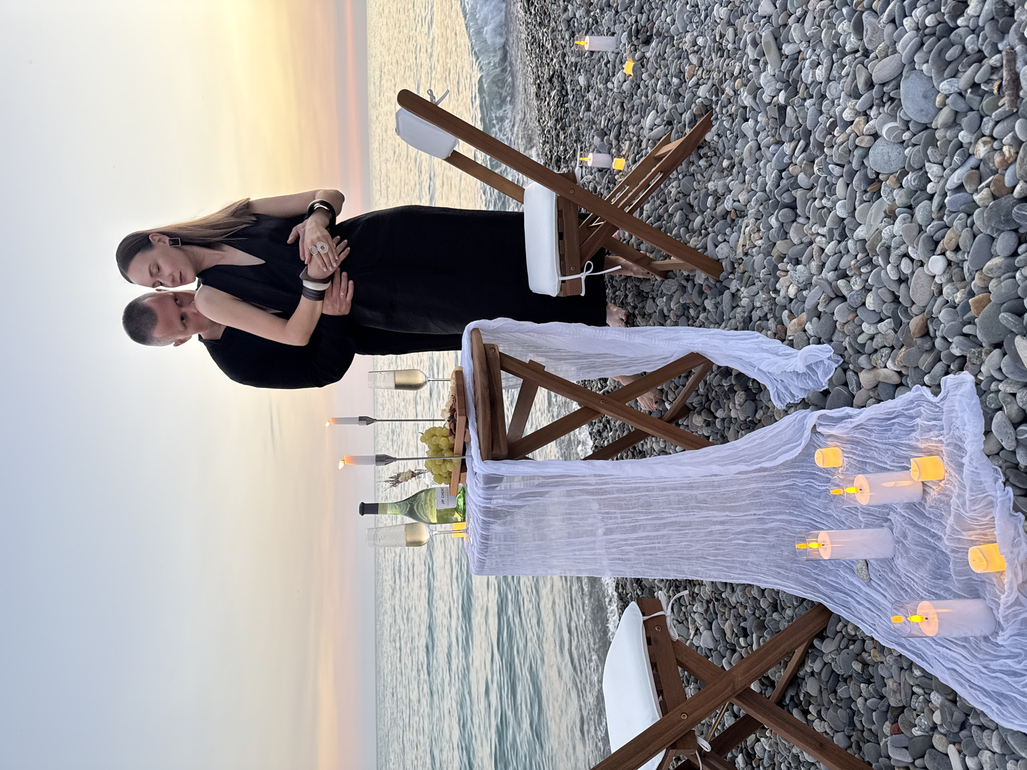 A man and a woman embrace on a pebble beach at sunset. The man, wearing a black shirt, has his arms around the woman, who is in a black dress. In the foreground, a small wooden table is set for a romantic meal with a white tablecloth, two glasses of champagne, a bottle of white wine, grapes, and a cheese board. Several lit candles are scattered around the table and chairs on the beach, creating a warm ambiance. The sea is visible in the background with gentle waves lapping at the shore. The sky displays soft hues of orange and pink from the setting sun. The scene appears to be set in Sochi, Russia, given the context provided.