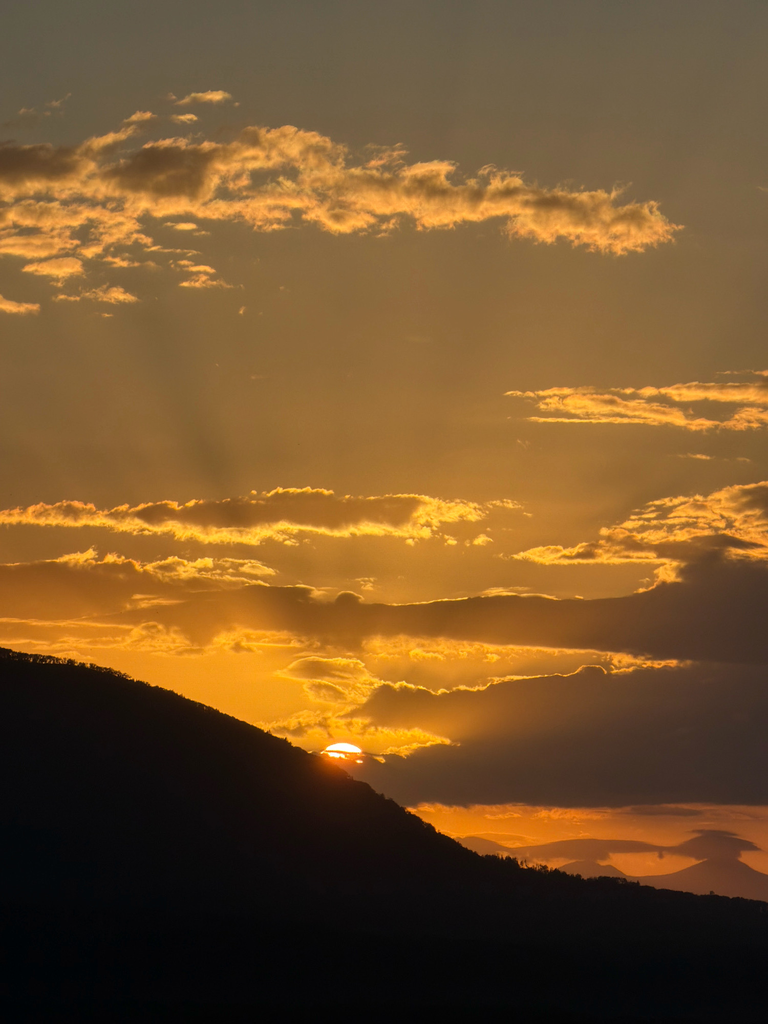The image captures a dramatic sunset over a mountainous landscape. The sky is a vibrant gradient of oranges and yellows, with scattered clouds catching the last rays of sunlight, creating a luminous effect. The sun itself is partially obscured by a dark, silhouetted mountain range in the foreground, creating a striking contrast. The texture of the clouds varies from wispy and ethereal to thick and dramatic. The foreground is dominated by the dark silhouette of a steep mountain slope, its peak lined with the faint outline of trees. Further in the distance, other mountain peaks are visible, their forms softened by the atmospheric haze and sunset light. The overall impression is one of natural beauty and the grandeur of a setting sun.