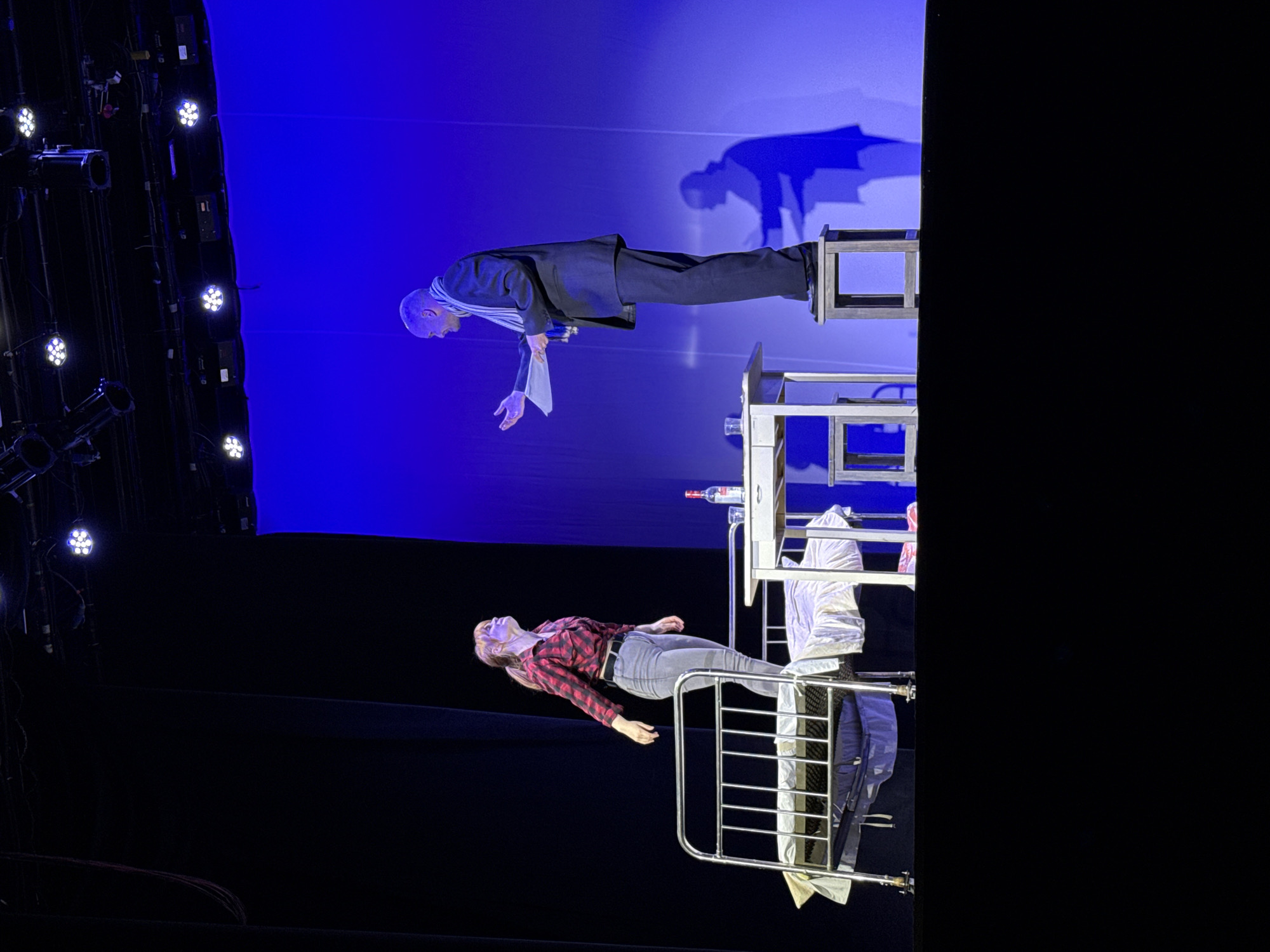 A dimly lit stage, likely inside a theater, depicts two people engaged in a theatrical performance.
At the top, a man dressed in a black suit with a white scarf lies horizontally, suspended between two small tables. His face is visible. A shadow of the man is visible on the blue backdrop behind him.
Below the man, a woman in a red and black plaid shirt and jeans lies horizontally on a metal bed frame. Her face is visible.
A table with a bottle on it stands near the floating man, and a white jacket hangs off of it.
The location is potentially Chaikovsky, Russia.