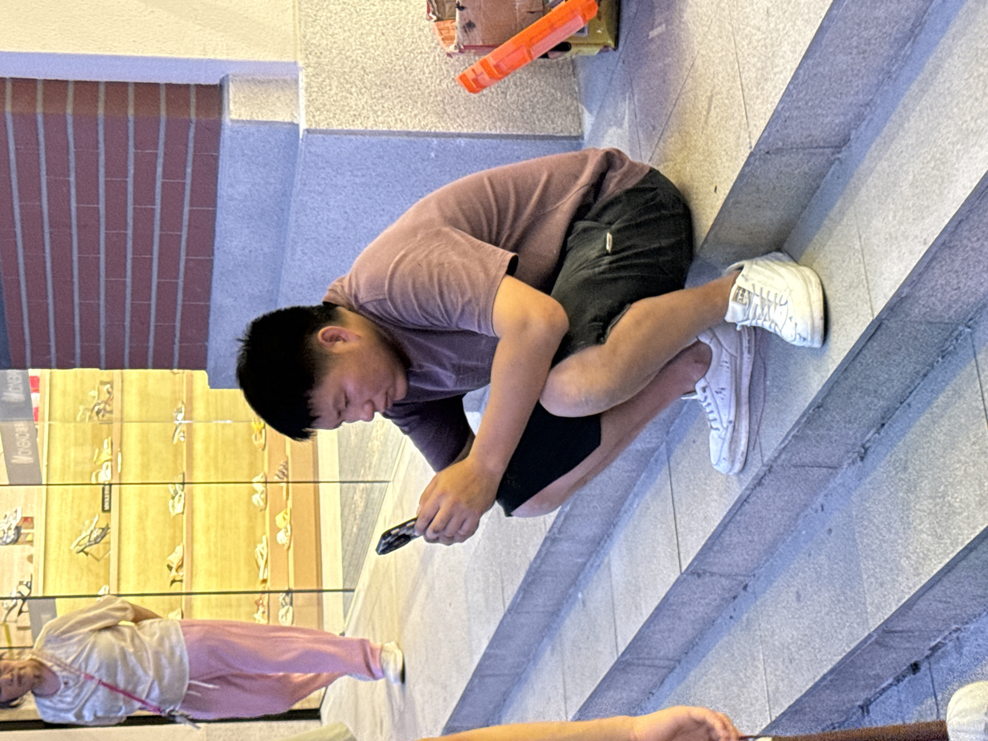 In Yuzhong District, China, a man of East Asian descent is crouched on a set of concrete stairs, looking down at a smartphone he holds in his hands. He is wearing a short-sleeved, muted purple t-shirt and dark shorts. His white sneakers are visible on his feet. To his right, a cardboard box with an orange object protruding from it sits on the ground. In the background, a woman in pink pants and a light-colored shirt walks past a storefront displaying shoes on shelves. The storefront appears to be illuminated by warm light. The architecture of the building features a brick facade above the stairs.