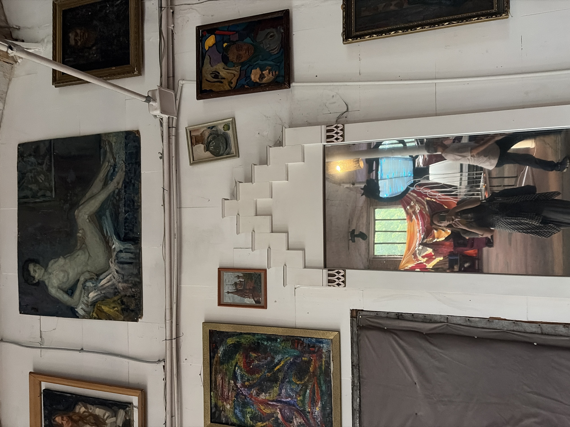 The image shows a wall with several framed paintings and a large mirror reflecting a room. The paintings are of various subjects and styles. On the top row, there is a painting of a reclining nude, a painting of a landscape with a church, and a dark portrait. In the middle row, there is an abstract painting, a still life with vases, and a painting of two faces with abstract elements. On the bottom, there is a large framed mirror that reflects a person taking a photo of themselves, a man walking, and a room with colorful fabric decorations and large windows. The wall appears to be white and textured, with some electrical conduits and wires running across it. The overall impression is that of an art studio or a gallery space. The city of Il'inskiy Pogost, Russia, is mentioned, but there are no specific visual cues in the image to confirm this location.
