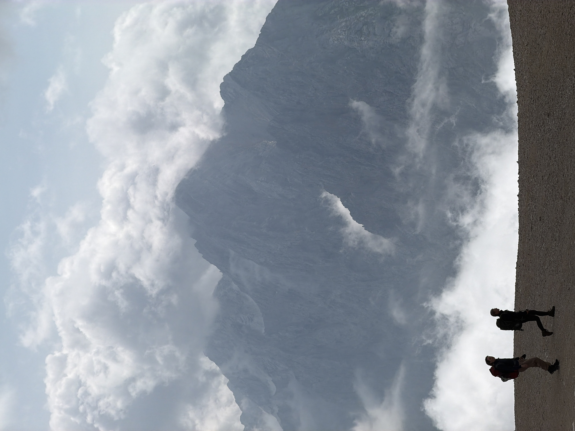 The image depicts two hikers with backpacks silhouetted against a dramatic mountain landscape. The hikers are walking on a gravelly ridge in the foreground. Behind them, a jagged, mist-shrouded mountain range dominates the background, with clouds swirling around its peaks. The sky above is a pale blue with scattered white clouds. The scene suggests a challenging and breathtaking hiking experience, likely in a mountainous region. While the image captures a natural landscape, there is no specific information in the visual content to confirm it is located in Garmisch-Partenkirchen, Germany. The hikers appear to be moving away from the viewer, and their interaction is limited to their shared presence in the environment.