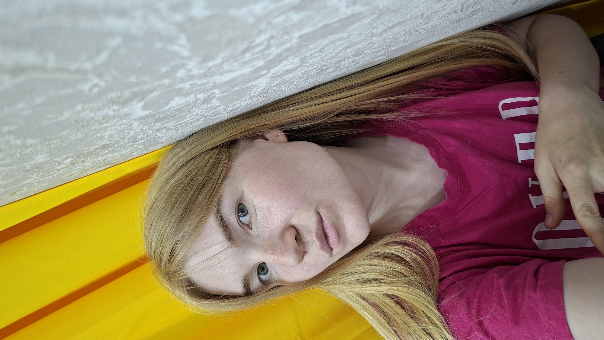 A young woman with long, blonde hair and freckles on her face is leaning against a textured wall. She is wearing a bright pink t-shirt with white lettering. To her left, a bright yellow curtain is visible. The scene appears to be indoors. The image was taken in the city of Chaikovsky, Russia.