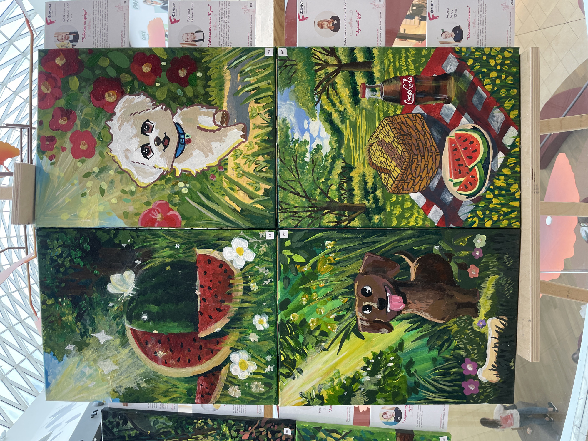 The image shows a collection of four acrylic paintings displayed on easels, likely at an art exhibition or gallery. The paintings are presented in a two-by-two grid.
The top left painting depicts a whole and sliced watermelon in a sunlit grassy area with a white butterfly. The top right painting features a fluffy white dog sitting among red flowers. The bottom left painting shows a brown dachshund in a sun-dappled forest setting with a bone. The bottom right painting illustrates a picnic scene with a basket, sliced watermelons, and a Coca-Cola bottle on a checkered blanket in a park-like setting.
On the right side of the displayed paintings are informational cards, each with a photograph of a child, a name, age, and a title of their artwork in Russian. These cards are positioned to the right of the paintings they correspond to, suggesting an exhibition of children's art.
The background indicates an indoor environment with a distinctive ceiling structure made of geometric glass panels, possibly a modern building or shopping mall. Based on the context of an art exhibition featuring children's works with Russian text on the informational cards, it is highly probable that this scene is located in the city of Perm, Russia. The overall impression is a vibrant display of artwork, likely a public or community event.