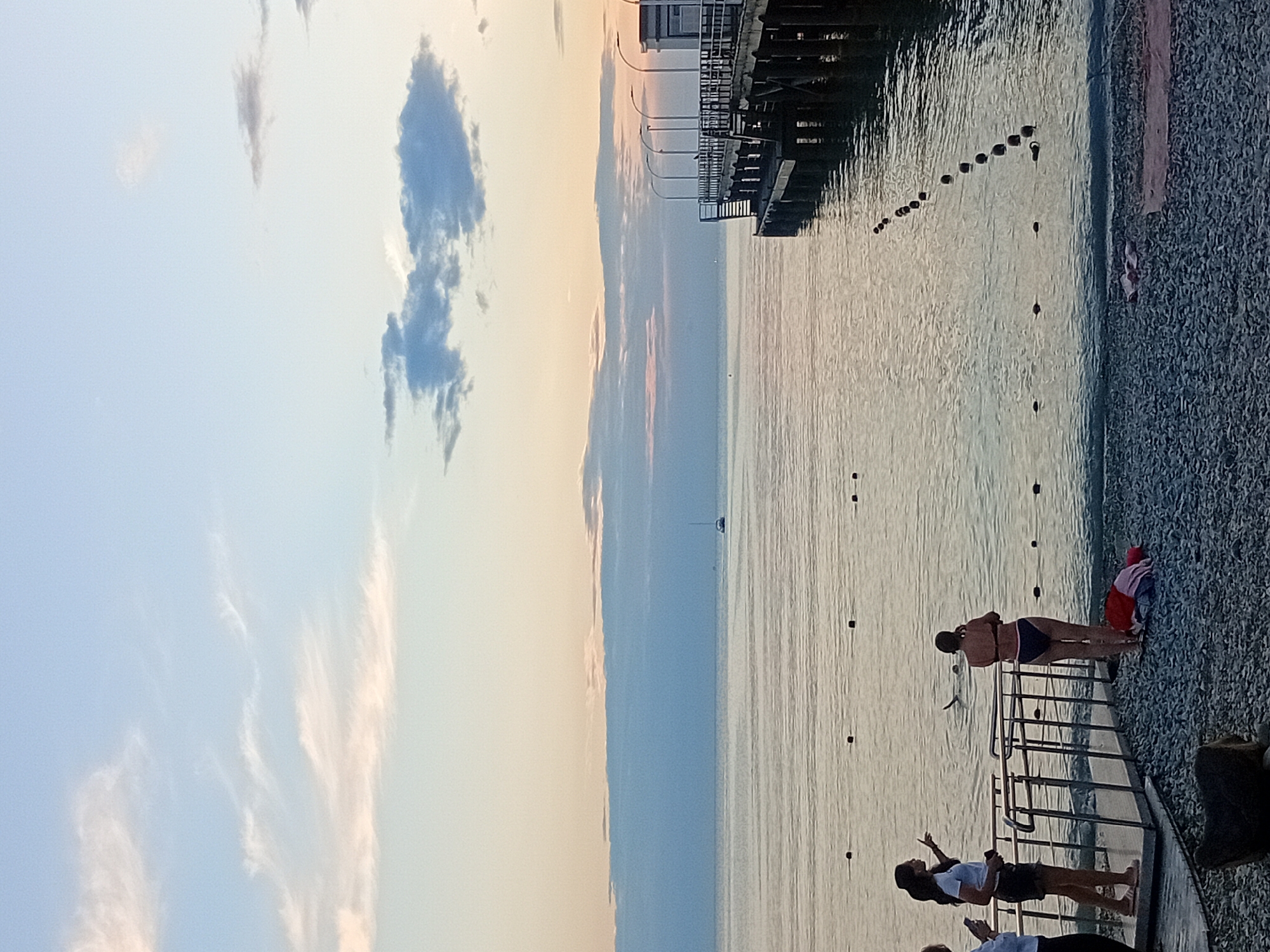 The image depicts a calm sea at dusk, with soft, pastel-colored clouds in the sky. A wooden pier extends from the right side of the frame into the water, featuring lights along its railing. On the left side, a paved walkway leads to the water's edge, where two women are standing. One woman, wearing a white t-shirt and shorts, points towards the sea, while the other, in a black bikini, stands facing away from the camera. In the water near them, another person appears to be swimming or floating. The shore is covered in pebbles. In the distance, a sailboat is visible on the horizon. The scene suggests a peaceful evening at a seaside location, likely in Novorossiysk, Russia, given the context of the query.
