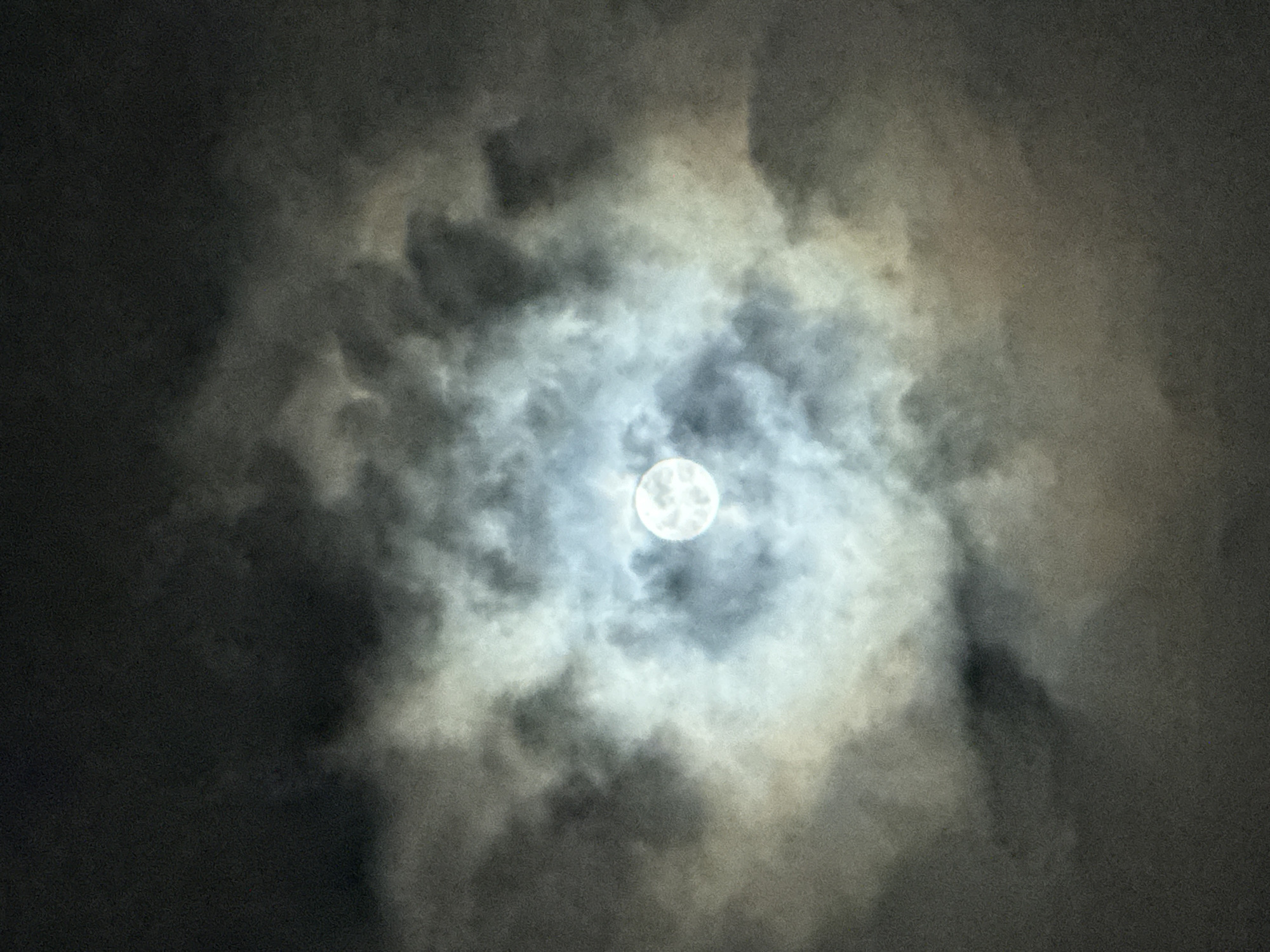 A full moon is partially obscured by clouds. The moon is positioned in the center of the frame, emitting a bright, diffused light that illuminates the surrounding clouds. The clouds are varied in texture and density, with some appearing wispy and translucent, while others are thicker and darker, creating a dramatic contrast with the moonlight. The overall color palette is dominated by shades of gray, white, and a subtle hint of yellow or brown in the clouds. The background is a deep, dark sky. There are no people, buildings, or identifiable landmarks visible in the image, thus it is impossible to determine the location within the city of Nha Trang, Vietnam.