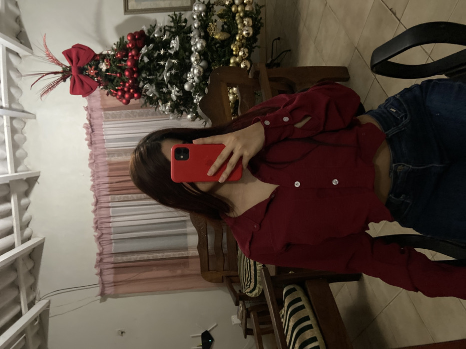 A young woman in a red crop-top blouse and blue jeans takes a mirror selfie, obscuring her face with a red phone. Behind her, a decorated Christmas tree stands near a window with pink curtains. Wooden furniture and a black office chair are visible. The setting appears to be a home interior. The room is illuminated by soft, ambient lighting.