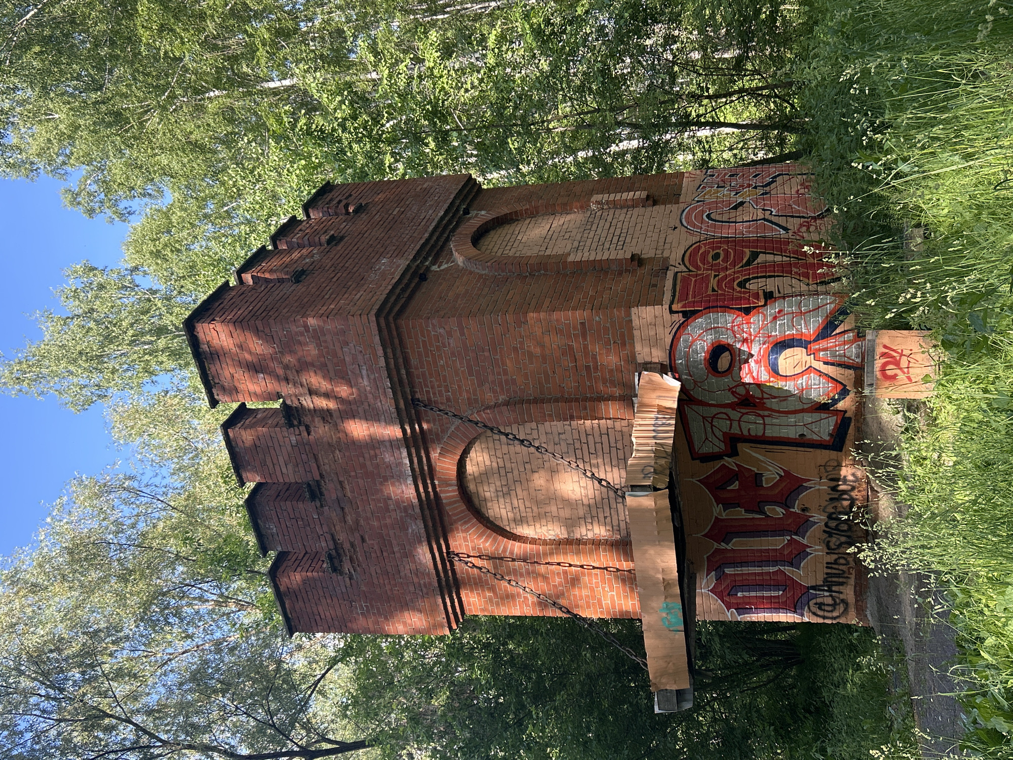 A tall, red brick structure resembling a small turret or watchtower stands in a wooded area. The structure has a crenellated top and arched openings that appear to be boarded up with light-colored material. Heavy chains are attached to the sides of the structure, suspending a makeshift platform made of cardboard and wood. The lower portion of the structure is covered in graffiti with large, stylized letters in red, gray, and black. The area around the structure is overgrown with tall green grass and wildflowers. In the background, tall green trees with bright green leaves create a dense canopy. The sky above is clear and blue. The structure is located in a park area in Chaikovsky, Russia.