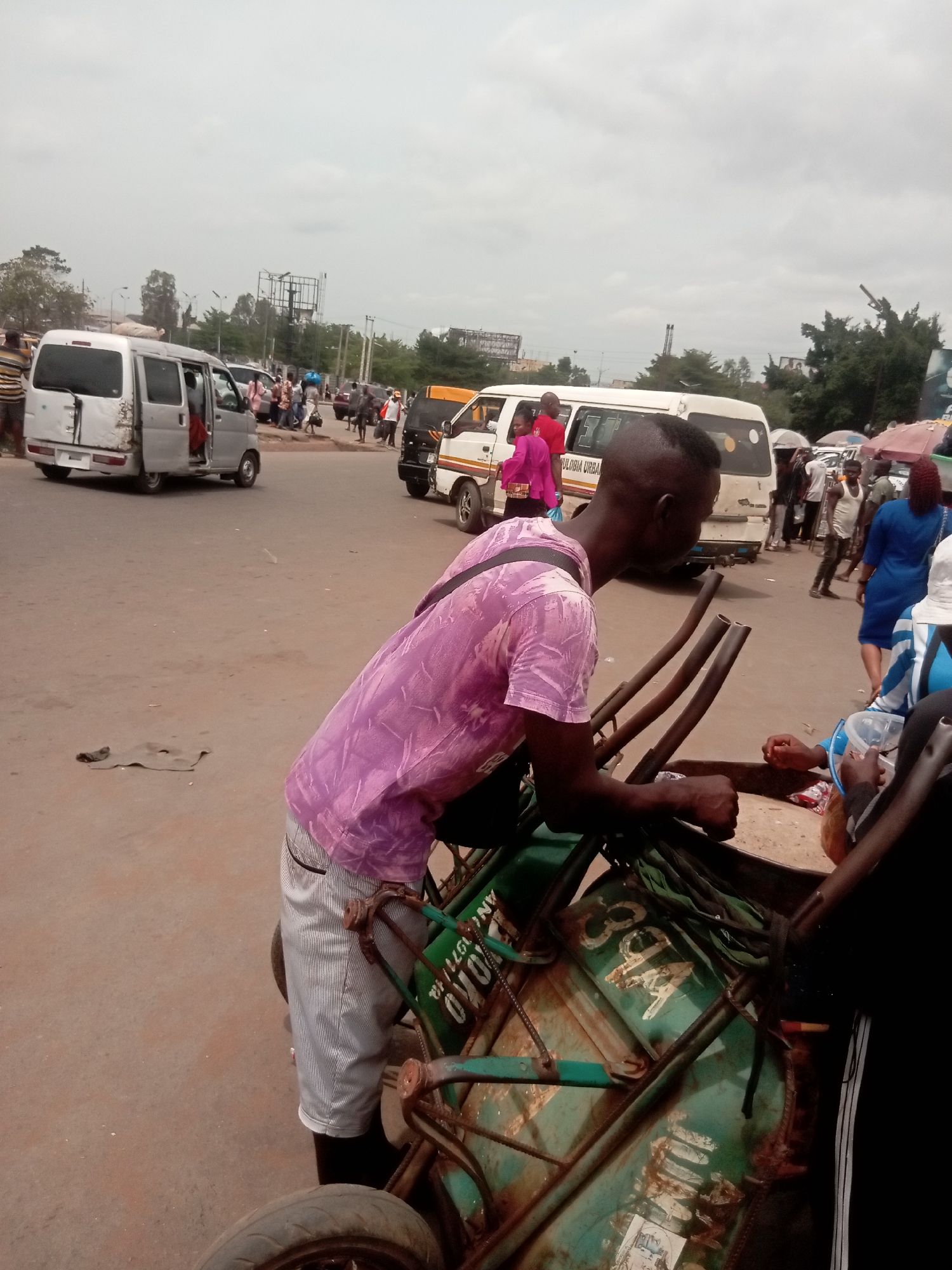 The image depicts a street scene in Onitsha, Nigeria. A man in a pink shirt and striped shorts is leaning over a hand-pushed cart, possibly interacting with someone out of frame. The cart is green and appears to be quite worn, with visible lettering "OYO" and "ANL. 1071 HR." Several vehicles, including vans, are visible in the background, along with pedestrians on the street. The sky is overcast. The general environment appears to be a busy intersection or roadside area, typical of urban street life in Onitsha.