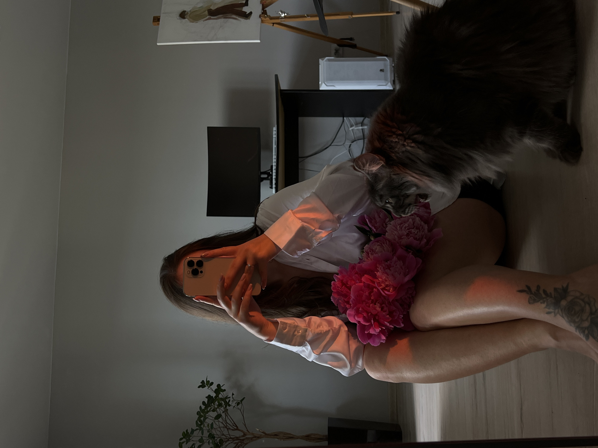 A young woman with long brown hair is sitting on the floor, holding a bouquet of pink peonies and taking a selfie with her phone. She is wearing a white button-down shirt and black shorts. To her right, a large, fluffy grey cat is lying down, looking towards the woman. In the background, a desk with a computer monitor is visible, along with a painting on an easel. The scene appears to be indoors, in a room with neutral-colored walls and a wooden floor. The lighting suggests it is late afternoon or early evening, with warm light casting shadows.