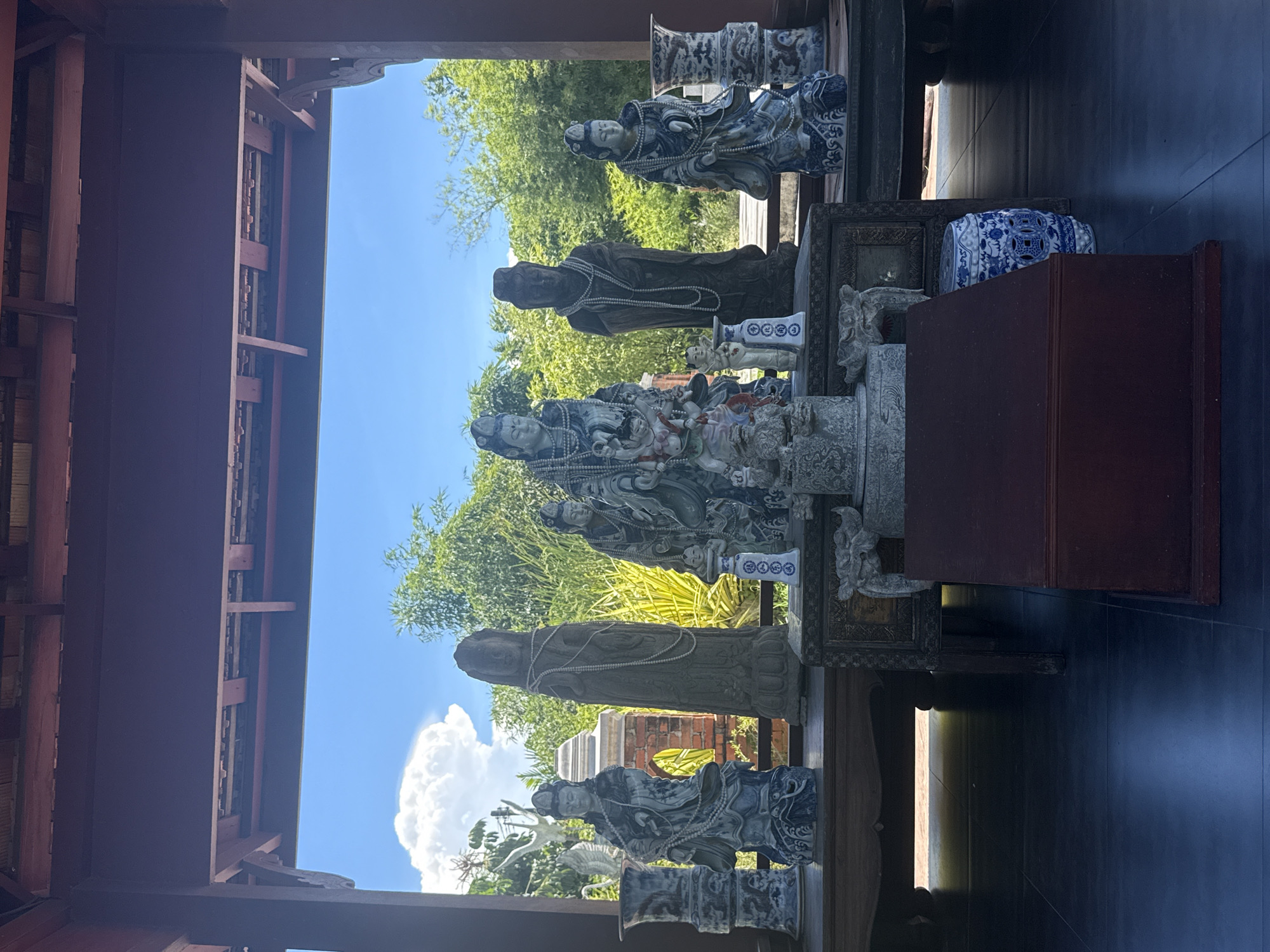 The image depicts a collection of statues displayed indoors, likely in a building located in Bang Pu Subdistrict Municipality, Thailand. The statues, varying in style and material (possibly porcelain and stone), are arranged along a ledge or shelf. Behind the statues, an outdoor scene is visible through an opening, showing trees, a building with arched brickwork, and a bright blue sky with puffy white clouds. The indoor space features dark wood beams and supports, suggesting traditional architecture. The statues are the primary subjects, with the external landscape serving as a backdrop. The floor is a dark, glossy surface.