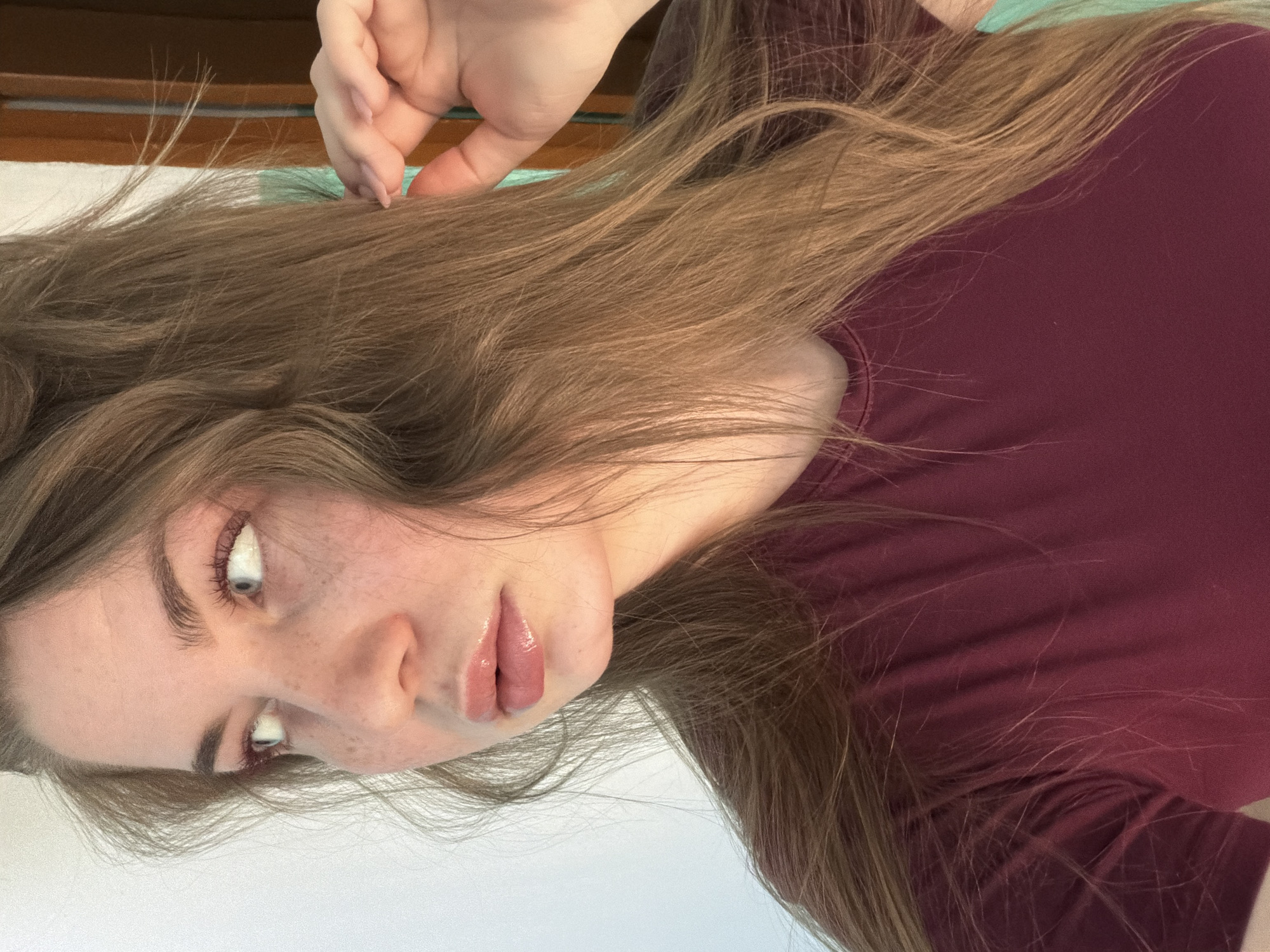 A woman with long brown hair, freckles, and light eyes is visible. She is wearing a maroon-colored shirt. Her right hand is near her hair. The background is indistinct. The location is not discernible.