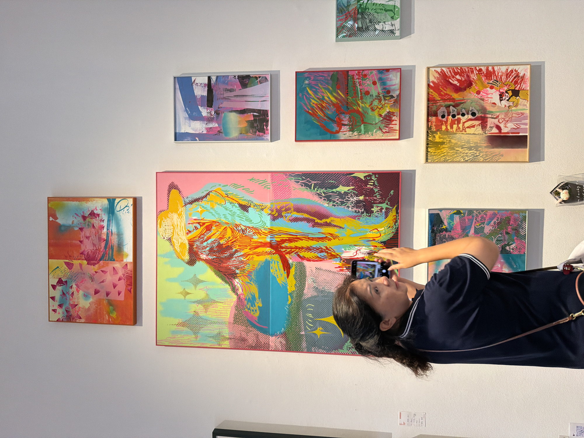 The image captures a detailed scene within an art gallery located in the Chongqing High-tech Zone, China. An adult East Asian female, with dark hair pulled back, is positioned with her back mostly to the viewer, angled slightly to her right. She wears a dark blue polo shirt with white stripes on the collar and sleeve cuffs and a brown cross-body bag strap across her shoulder. With both hands, she holds a black smartphone horizontally, pointing it towards a large, vibrant artwork displayed on the white wall. Her gaze is directed at the phone's screen, which reflects the artwork.
The wall is adorned with multiple colorful, contemporary abstract paintings of various sizes and orientations. The prominent central artwork, being photographed by the woman, is a large vertical canvas depicting a stylized figure or form rendered in bold, contrasting hues of yellow, orange, red, blue, and pink against a patterned background. Above this, a horizontal painting features warm pink, orange, and red tones, with hints of blue and geometric triangular shapes. To the right of the central piece, a vertical stack of three smaller paintings is visible: the top one featuring abstract blue, pink, and black strokes; the middle one framed in red, showcasing dynamic red, yellow, and green elements; and the bottom one, a smaller square, with green, black, and white patterns. Below the central artwork, a square painting displays strong red and yellow brushstrokes, possibly including a small cartoonish figure and circular elements. A small, wrapped item with a white bunny ornament sits on a low display next to this bottom-right painting. On the far left, a white piece of paper with Chinese characters and a QR code is affixed to the wall. The gallery space features clean white walls and bright lighting.