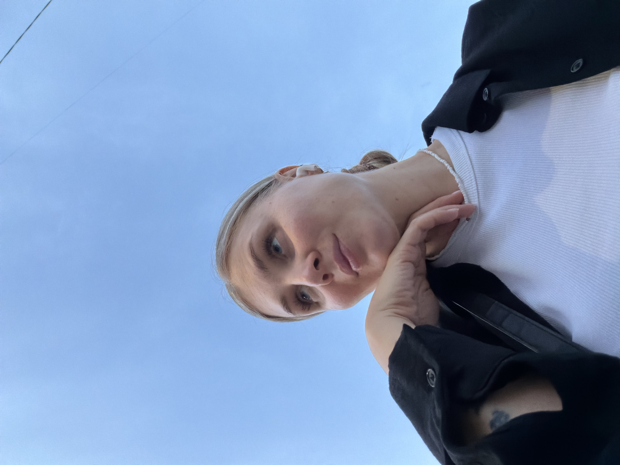 The image shows a fair-skinned, blonde woman with blue eyes, looking down at the camera. She is wearing a white shirt, a black jacket, and a white beaded necklace. She is resting her chin on her left hand. The background is a clear, light blue sky. The location is in the city of Chaikovsky, Russia.