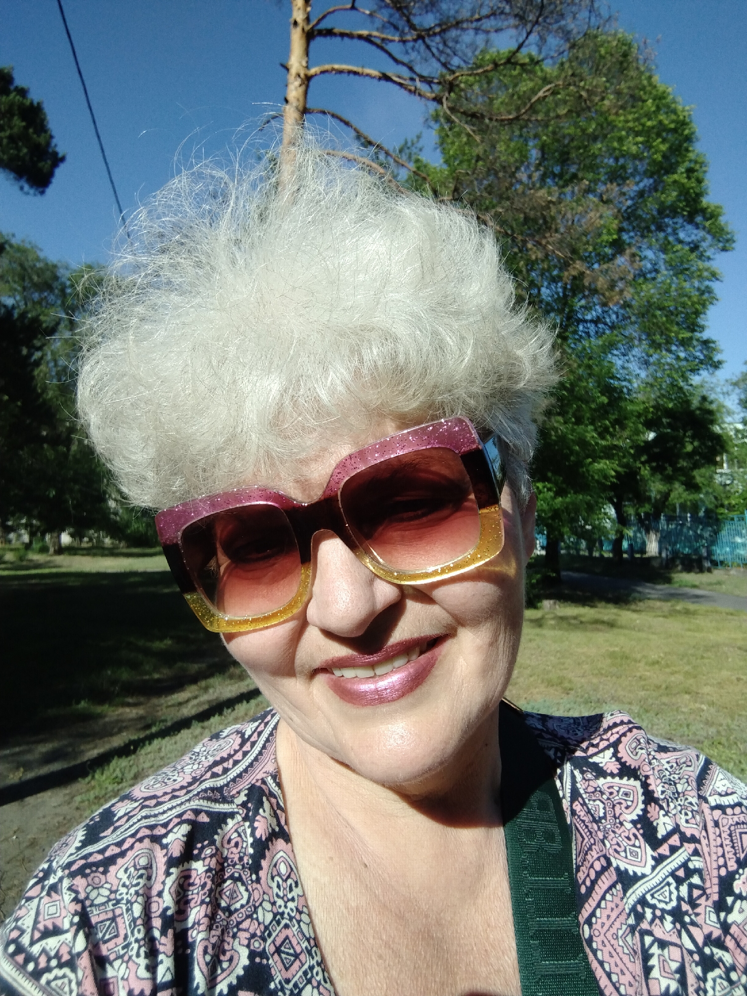 A woman with short, voluminous, white-grey hair smiles directly at the camera in what appears to be an outdoor selfie taken in Horishni Plavni, Ukraine. She wears large, oversized sunglasses with glittery frames that are pink on top and yellow on the bottom, obscuring her eyes. Her lips are painted with a shimmery pink lipstick, and her smile reveals her teeth.
She is dressed in a patterned top or jacket featuring a design in dark blue, pink, and white. A plain, patterned green strap, possibly from a bag, is visible over her right shoulder.
The background shows a bright, sunny daytime scene with a clear blue sky. Trees with green leaves and some with visible branches are present. Sunlight casts shadows on the ground, which appears to be a mix of grass and dirt. A paved path and a light blue fence are visible in the middle distance, suggesting a park or public green space. The overall atmosphere conveyed is cheerful and relaxed. There is no legible text visible on any of the objects or in the background.