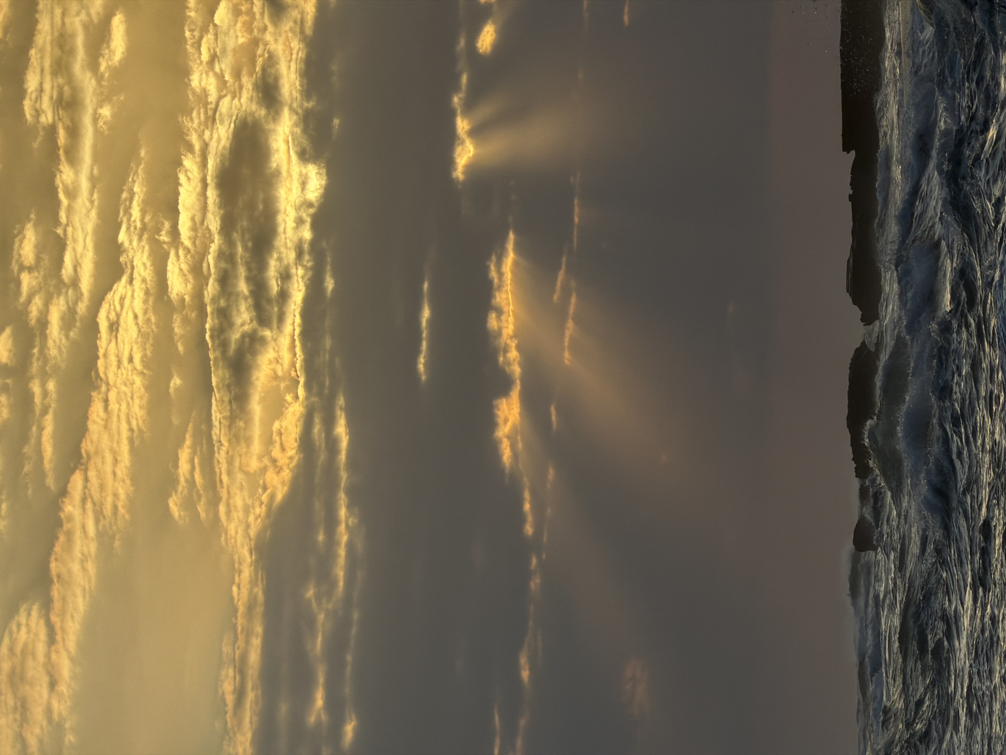 The image shows a rough sea with waves crashing against rocky outcrops. The sky is filled with dramatic clouds, illuminated by the golden rays of the sun peeking through. The overall mood is one of raw, natural power and beauty. The location is the city of Mengwi, Indonesia. There are no people or specific objects in the scene besides the natural elements of the sea, rocks, and sky.