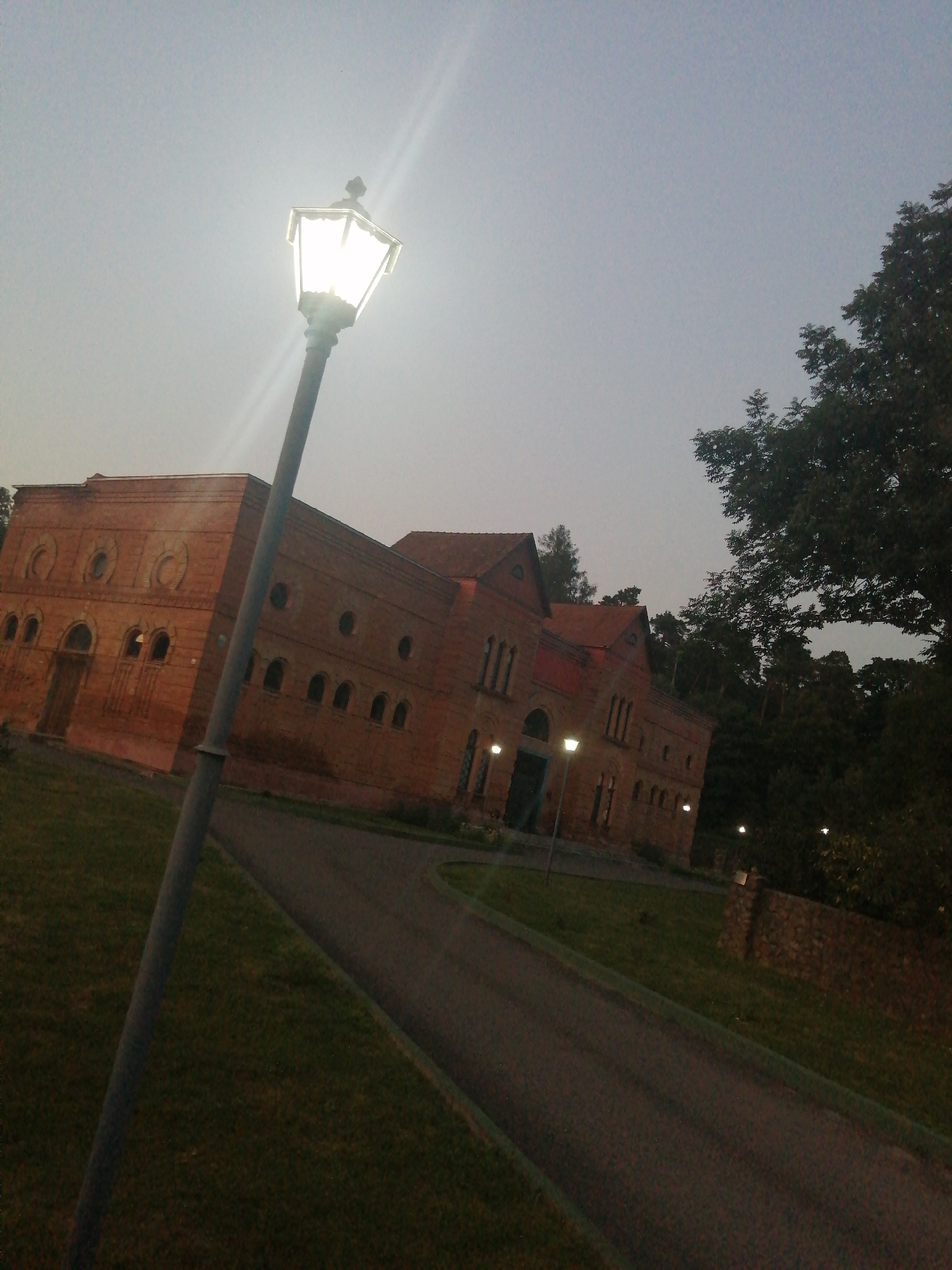 The image shows an outdoor scene at dusk. In the foreground, a tall, ornate street lamp is lit, casting a bright glow and creating lens flare. The lamp post is dark gray and positioned to the left of the frame. A paved pathway curves away from the viewer towards the right, bordered by a strip of green grass on the left and a stone wall on the right.
In the middle ground, a long, multi-story building constructed of reddish-brown brick is visible. The building has a distinctive architectural style with arched windows and decorative circular elements above some of the windows. The building appears to be a public or institutional structure. There are also several smaller, lit street lamps along the pathway in front of the building.
The background features a dense line of dark green trees, indicating a wooded area. The sky is a pale, overcast gray, suggesting twilight. The overall impression is a serene, slightly somber atmosphere. This scene is likely located in the city of Slonim, Belarus, though specific landmarks are not identifiable from the image alone. There are no people or animals visible in the frame.