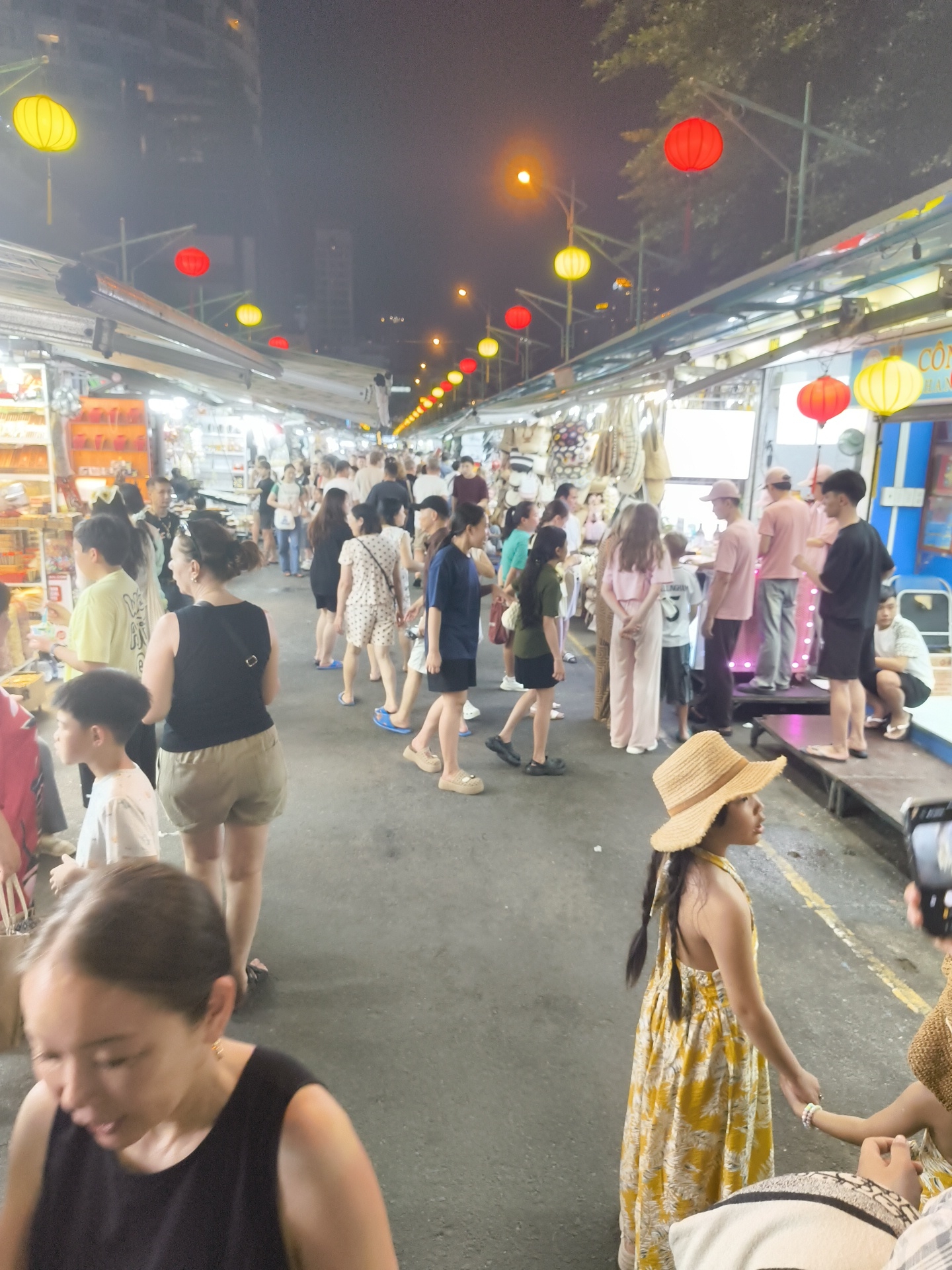 The image depicts a bustling night market in Nha Trang, Vietnam. Numerous people are browsing through stalls lining a street, illuminated by strings of red and yellow lanterns. The scene is vibrant and lively, suggesting a popular evening activity.
Several individuals are the focal point of the image. In the foreground, a woman in a black tank top and khaki shorts is visible, her face turned away from the camera. Further back, a young girl in a yellow floral dress and a straw hat stands holding someone's hand, looking to the side. Other shoppers are seen in various attire, walking and interacting within the market. The stalls appear to sell a variety of goods, with some displaying textiles and other items.
The time of day is clearly night, evidenced by the artificial lighting and the dark sky. The weather appears to be clear, as there are no signs of rain. The presence of lanterns, the types of stalls, and the overall atmosphere strongly suggest a market in Southeast Asia, consistent with the Nha Trang, Vietnam context. The general mood seems to be one of casual browsing and enjoyment, with the crowds indicating a successful and active market. There is no discernible text in the image.
