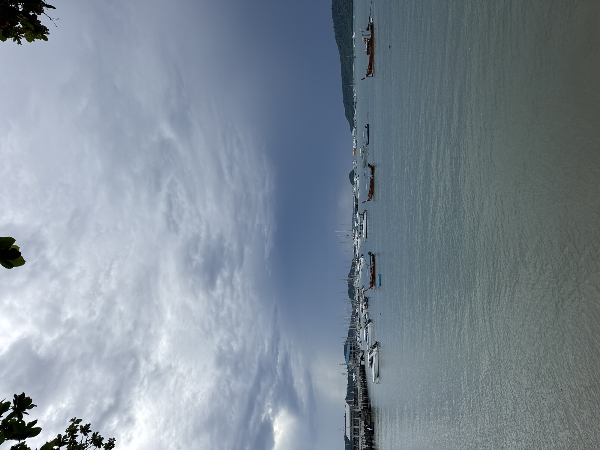 The image depicts a harbor in Rawai, Thailand, under a cloudy and dramatic sky. The water is a calm, greenish-blue, with small ripples on the surface. Numerous boats of various sizes are moored in the bay. Closer to the shore, there are several longtail boats with distinctive wooden hulls and overhead structures, as well as a few smaller white speedboats. Further out, a larger collection of white-sailed yachts and other vessels are visible, suggesting a busy marina. A long wooden pier extends into the water on the left side of the frame, with buildings and structures lining its edge. In the distance, a lush, green, hilly coastline forms the backdrop to the scene, with more boats scattered along its edge. The sky is a mix of light gray clouds and darker, more ominous storm clouds, with rays of sunlight breaking through in a few spots, creating a somewhat moody atmosphere. There are no people visible in the image, and no apparent actions or interactions taking place. The scene is serene, with the focus on the boats and the natural landscape.