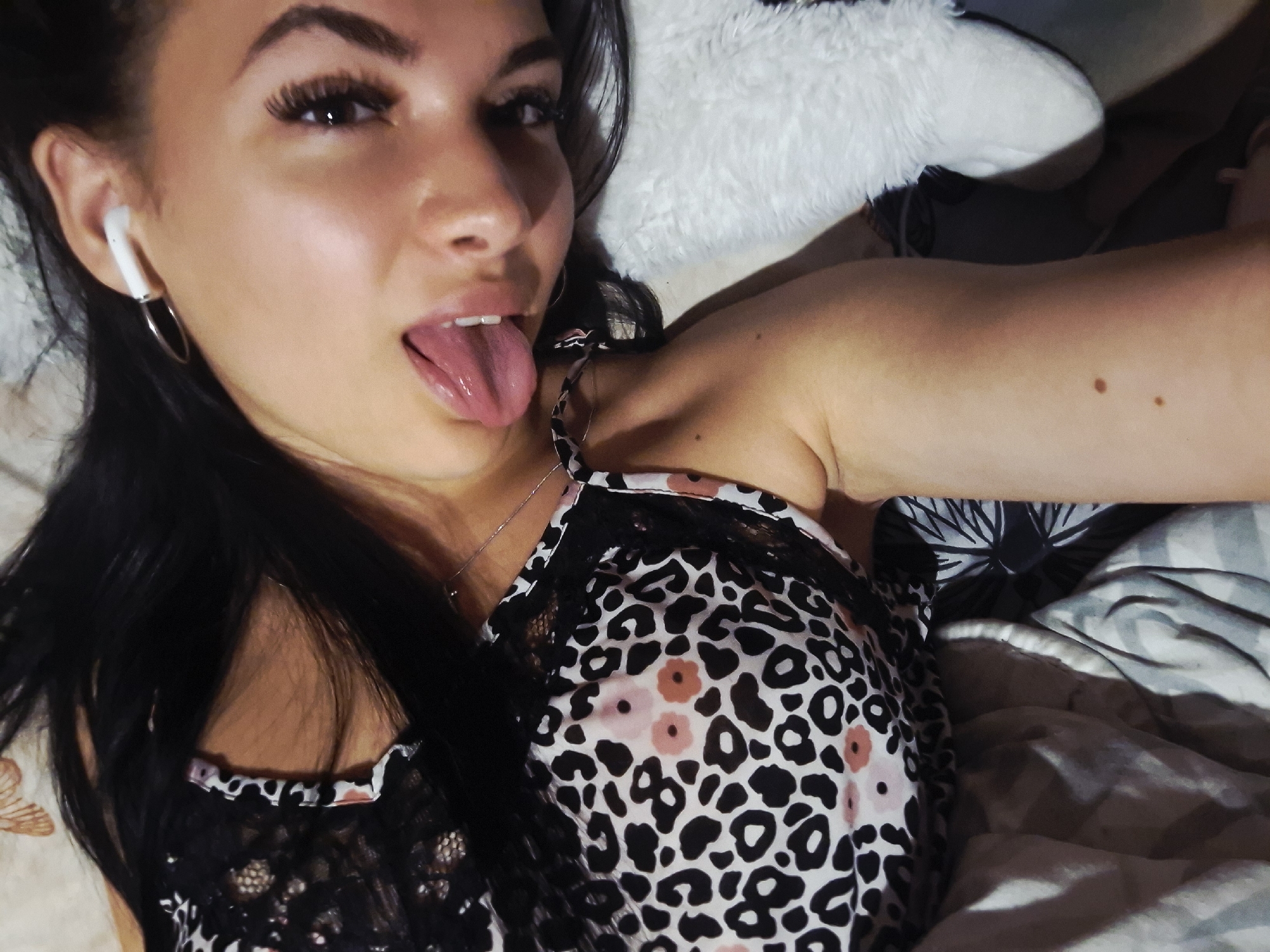 A young woman is captured in a close-up, high-angle shot, lying down indoors, possibly in a bed. She has long, dark hair and dark eyes, with prominent eyelashes. She is looking directly at the camera with a playful expression, her tongue sticking out.
She is wearing a white wireless earbud in her left ear, along with a silver hoop earring. A thin silver necklace is visible around her neck. Her attire includes a top with a distinctive pattern: black leopard spots on a white background, interspersed with some pink, flower-like shapes. The top also features black lace trim along the neckline and straps.
The background suggests a cozy indoor setting, likely a bedroom. To the left, a light-colored, possibly beige or cream, surface is visible. Behind her head and to the upper right, there is a soft, white, fluffy blanket or pillow. Further to the right, darker patterned fabric and a grey striped blanket or bedding are discernible. The lighting appears to be artificial or dim, highlighting the subject. No text is visible in the image. While the provided context suggests Krapets, Bulgaria, there are no visual cues in the image itself to confirm this location.