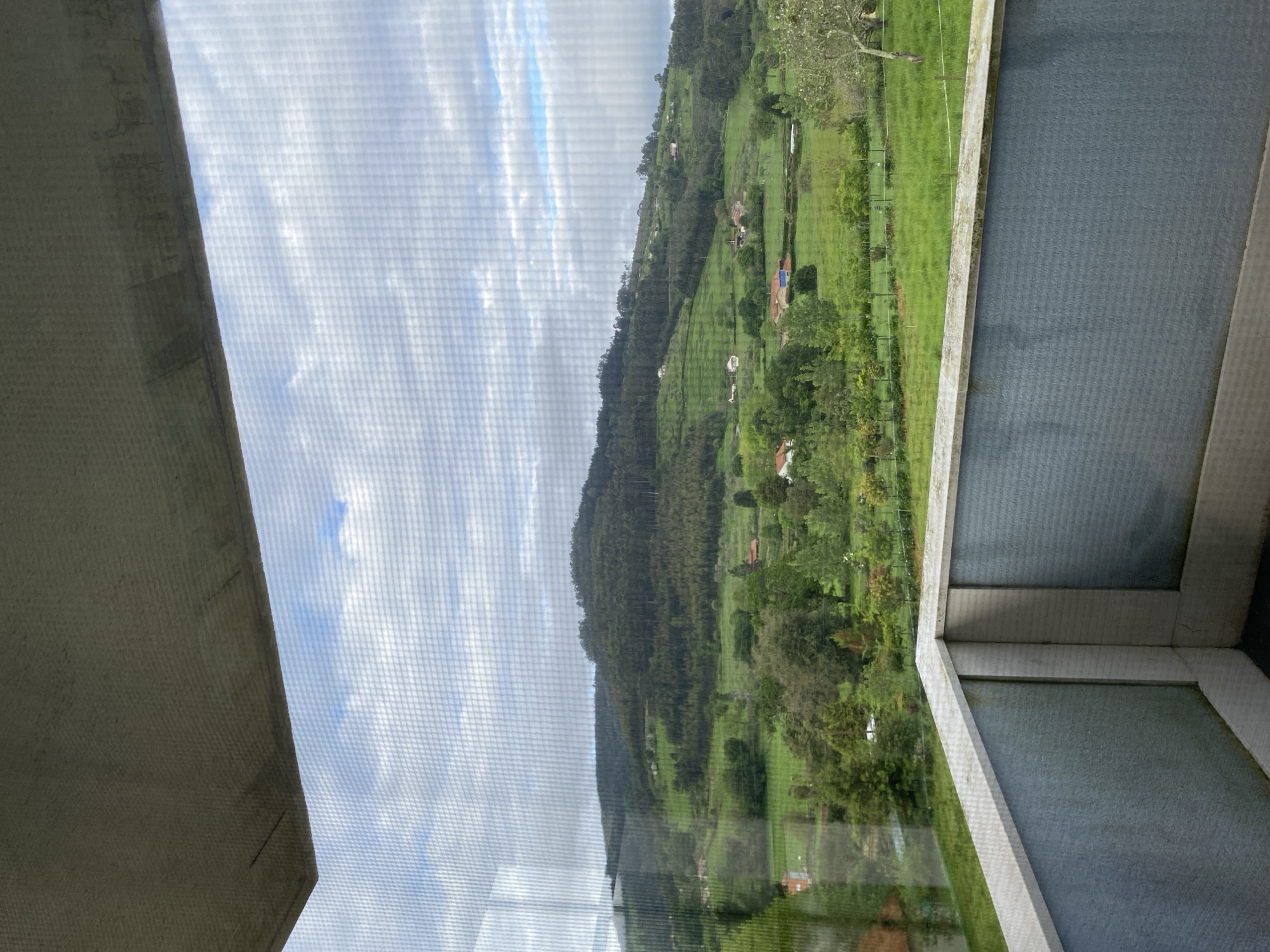A view from inside a building, possibly a residence, through a screened window, overlooking a hillside in what is presumed to be Villaviciosa, Spain. The landscape comprises of green fields, scattered trees, and a densely forested hill. There are a few small structures, likely houses, visible on the hillside. The sky is partially cloudy. The window frame and screen are prominent in the foreground, obscuring parts of the view. The photograph is slightly rotated counter-clockwise. No people or animals are visible in the image.