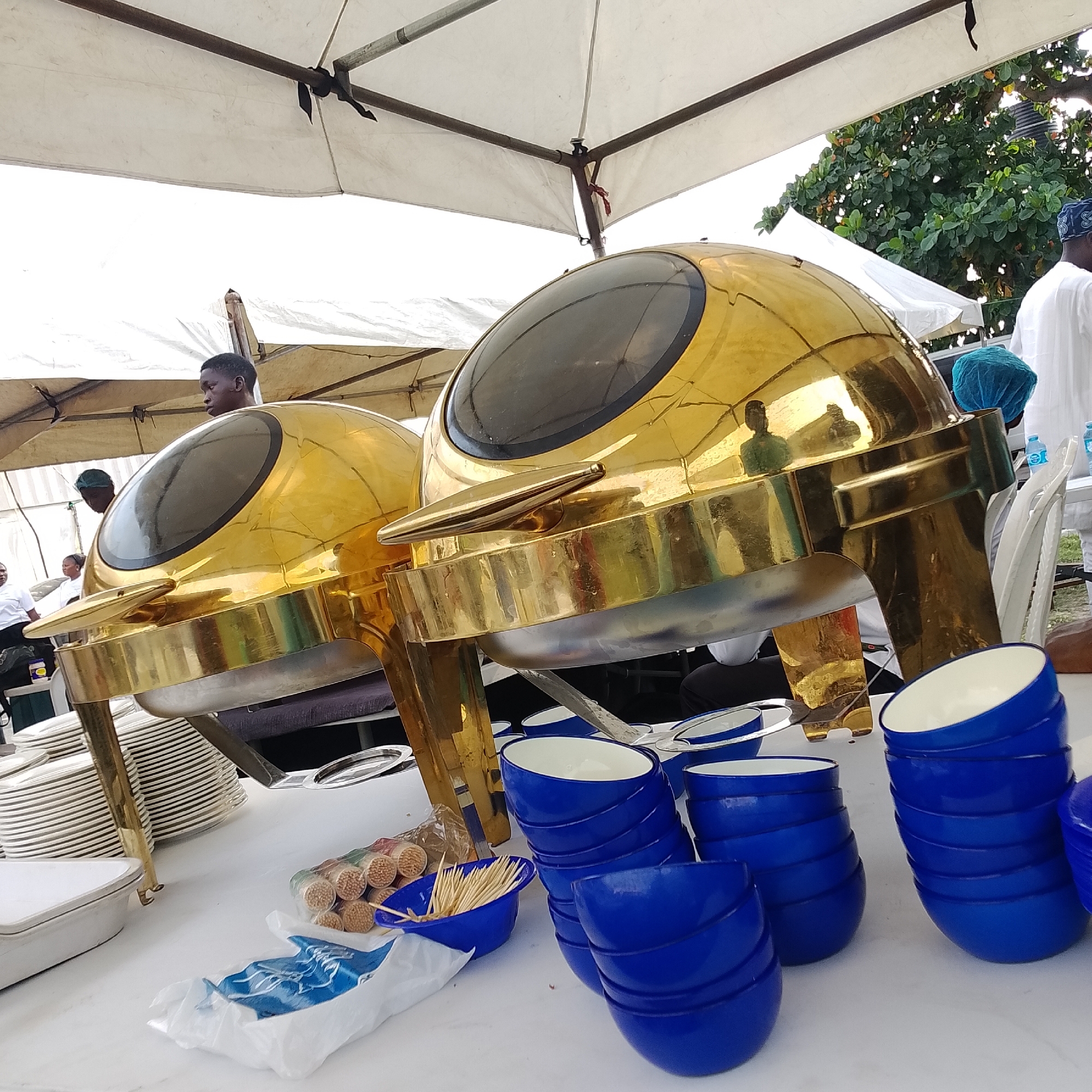 A close-up, low-angle outdoor shot shows two shiny gold chafing dishes with clear lids, likely at an event in Ikoyi, Nigeria. The chafing dishes are positioned on a white table laden with stacked white plates and other serving items. In the foreground, a pile of bright blue bowls is stacked, and next to them, a small blue bowl contains a bundle of wooden toothpicks. Behind the chafing dishes, a white tent provides shade. Several people, mostly wearing white, are visible in the background, some in the distance and one directly behind the right chafing dish, wearing a blue hair net. The overall scene suggests a catered event or food service setting.