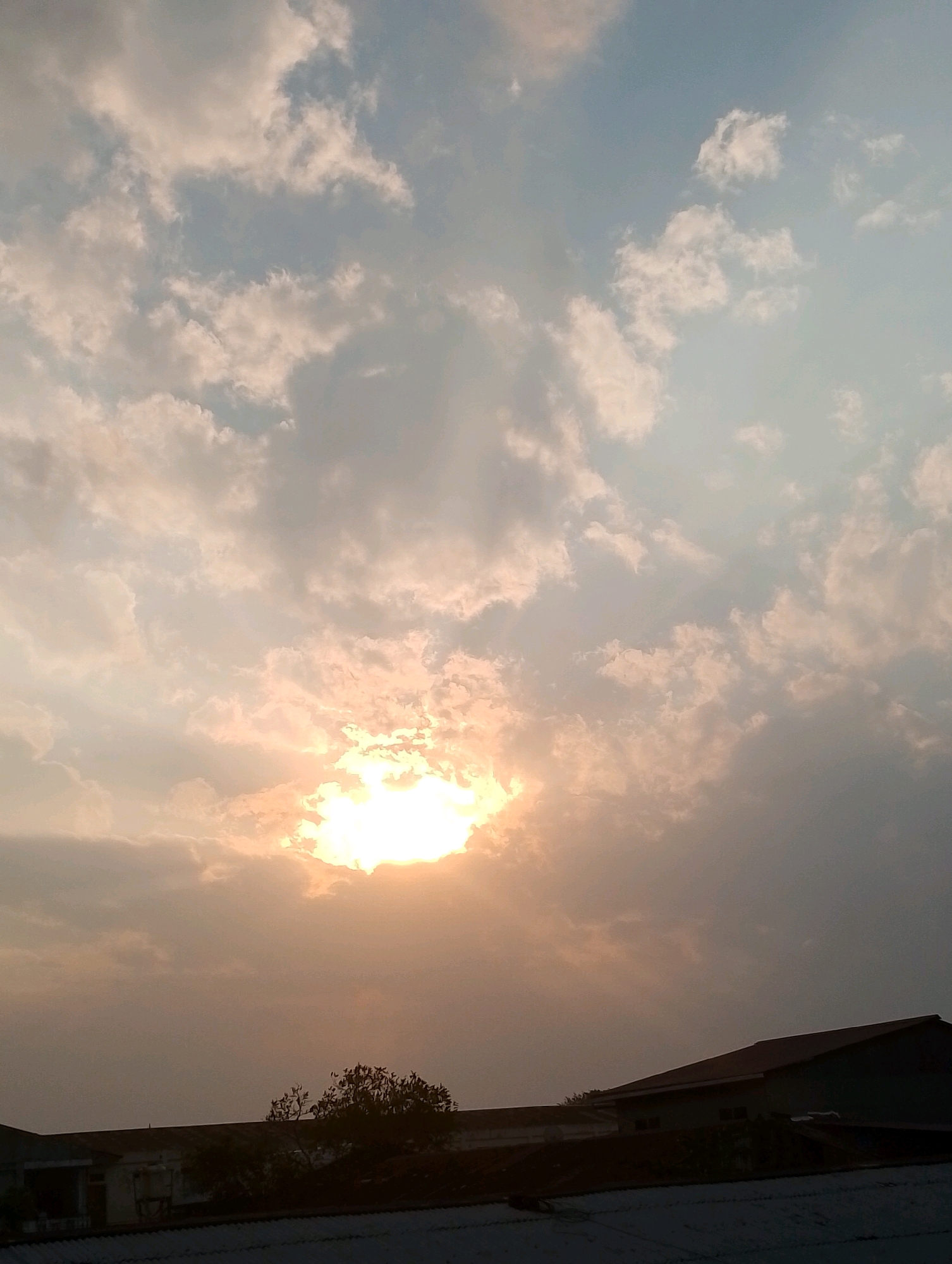 The image depicts a sky filled with scattered clouds, with the sun partially obscured by a dense cluster of them. The sun's rays are visible, casting a warm glow through the clouds, indicating either sunrise or sunset. Below the sky, the silhouettes of rooftops and trees are visible, suggesting an urban or suburban setting. The lower part of the image shows the roofs of several buildings, one with a red-tiled roof, and a dark silhouette of trees. The scene appears to be captured from an elevated perspective, looking towards the horizon. The time of day appears to be either early morning or late afternoon due to the lighting conditions. The overall atmosphere is serene and peaceful.