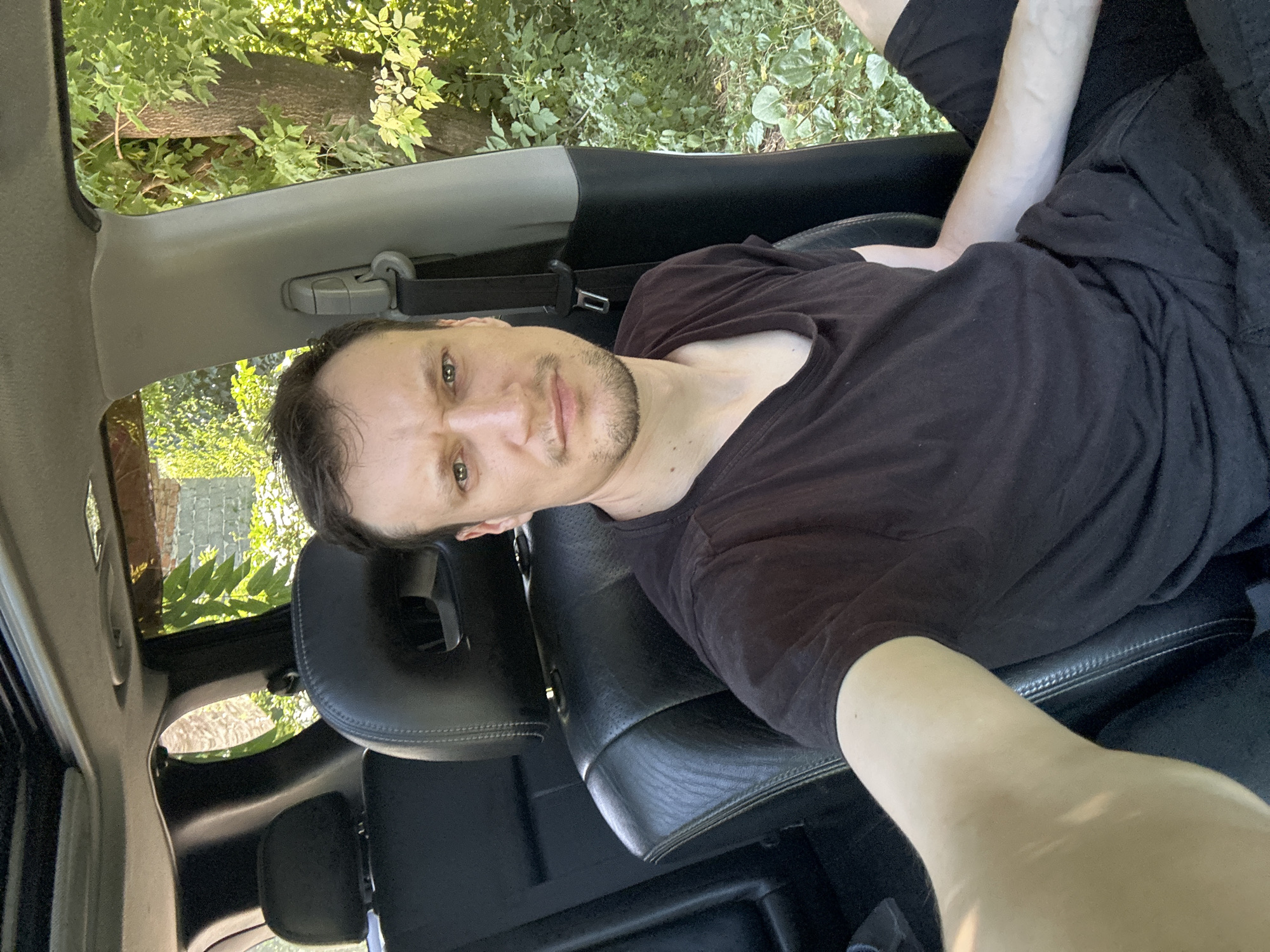 A man with short brown hair and a beard sits in the driver's seat of a car, facing the camera. He is wearing a dark t-shirt and dark shorts. His left arm is extended forward, holding the camera that took the photo. The car's interior, including the black leather seats and a sunroof, is visible. Outside the car, lush green foliage and trees suggest a rural or park-like setting. The overall impression is a selfie taken outdoors, possibly in a wooded area near Makiv, Ukraine.