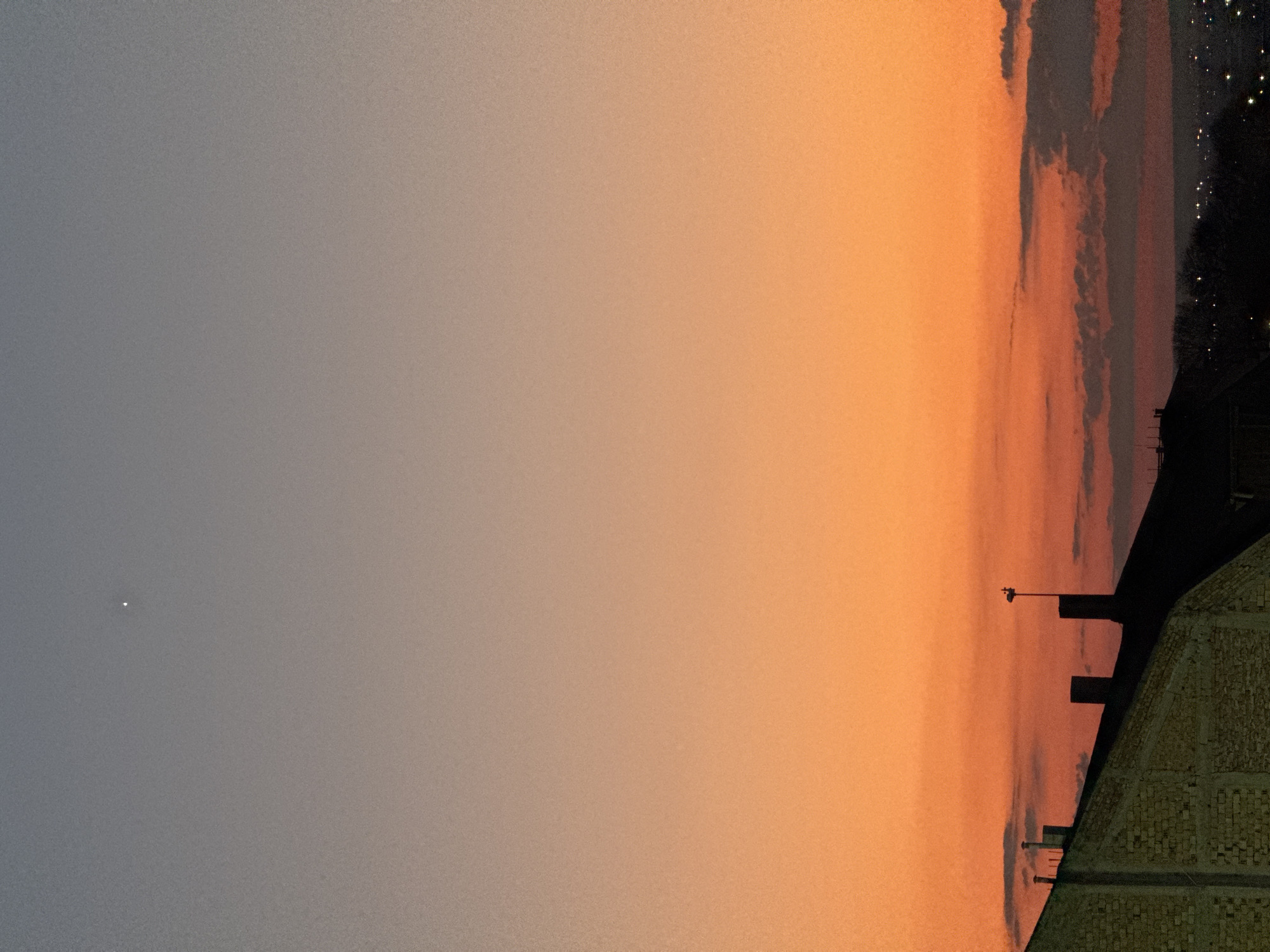 The image shows a view of the sky at dusk. The left side is a pale blue-gray, transitioning to orange on the right. A single bright star is visible on the left. On the right side, the silhouettes of buildings and trees are visible against the orange sky. The lights of the city can be seen in the distance to the top-right. The buildings include a brick structure, potentially the roof of a building, with a flagpole. The scene appears to be in an urban environment, likely Varna, Bulgaria.