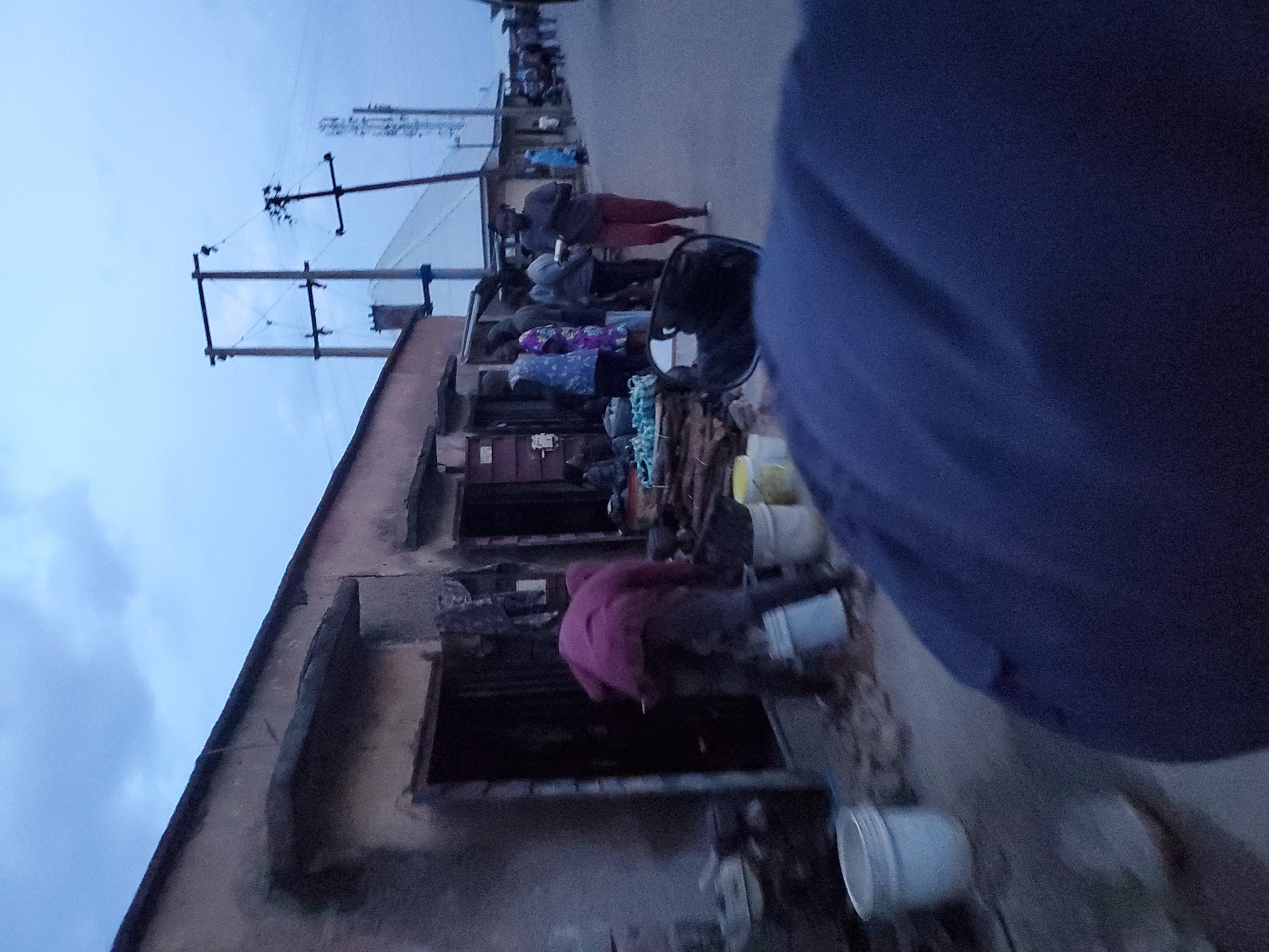The image depicts an outdoor scene in Jos, Nigeria, likely in the early evening given the dim lighting and overcast sky. The foreground is dominated by a dark blue fabric, possibly clothing or a blanket, obscuring much of the lower portion of the frame. To the left, a series of structures line a street. One building, a single-story structure with a weathered, textured facade, features open doorways and what appear to be small shops or stalls. A person in a pinkish-red jacket is bent over in front of one of these doorways, near several white and yellow buckets. There are also bundles of what could be ropes or textiles stacked on the ground.
Further down the street, several other people are visible, walking and standing. A woman in orange-red trousers and a dark top is holding something in her hands as she walks. Other individuals are seen in the background, some appearing to be vendors or shoppers. Utility poles with electrical wires crisscross the sky, and in the distance, a cell tower is faintly discernible. The overall atmosphere suggests a busy, perhaps commercial area, experiencing the transition from day to night. The road surface appears to be asphalt or a similar paved material.