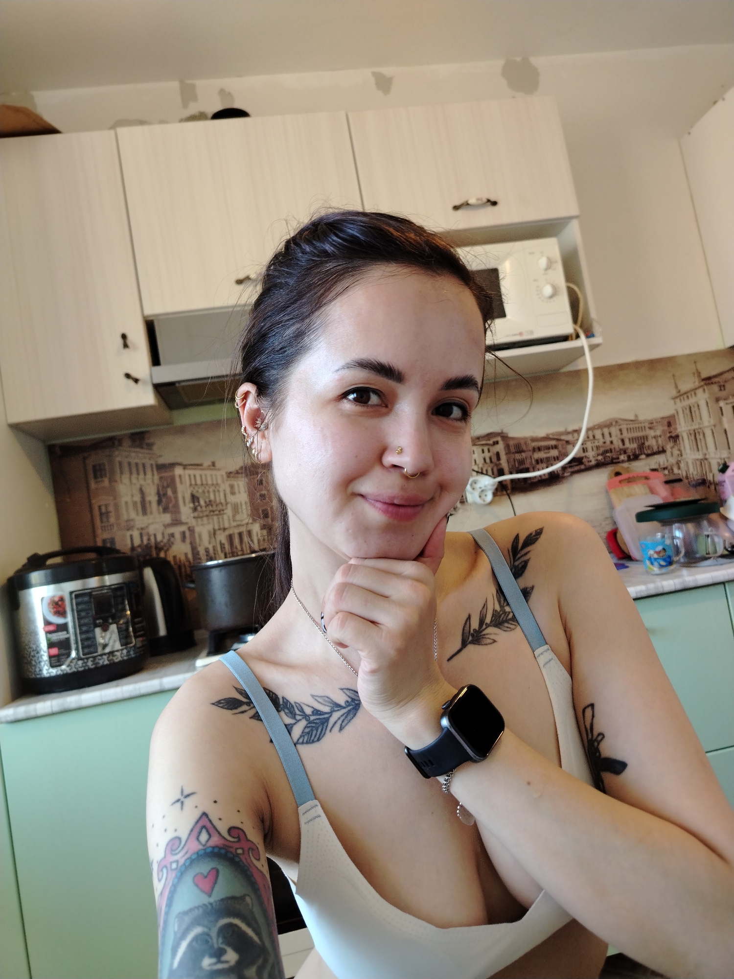 A medium-shot of a woman posing for a selfie in what appears to be a kitchen setting. The woman has dark hair pulled back, fair skin, and is wearing a white bra with blue straps. She has multiple piercings in her left ear and a nose ring. Her left arm is raised, with her hand resting under her chin. A black smartwatch is visible on her left wrist. The woman also has several tattoos on her arms and chest.
The background includes kitchen cabinets, a microwave, and a backsplash featuring a depiction of buildings in a European style. An electric pressure cooker and an electric kettle sit on the countertop. The location is not determinable from the image.