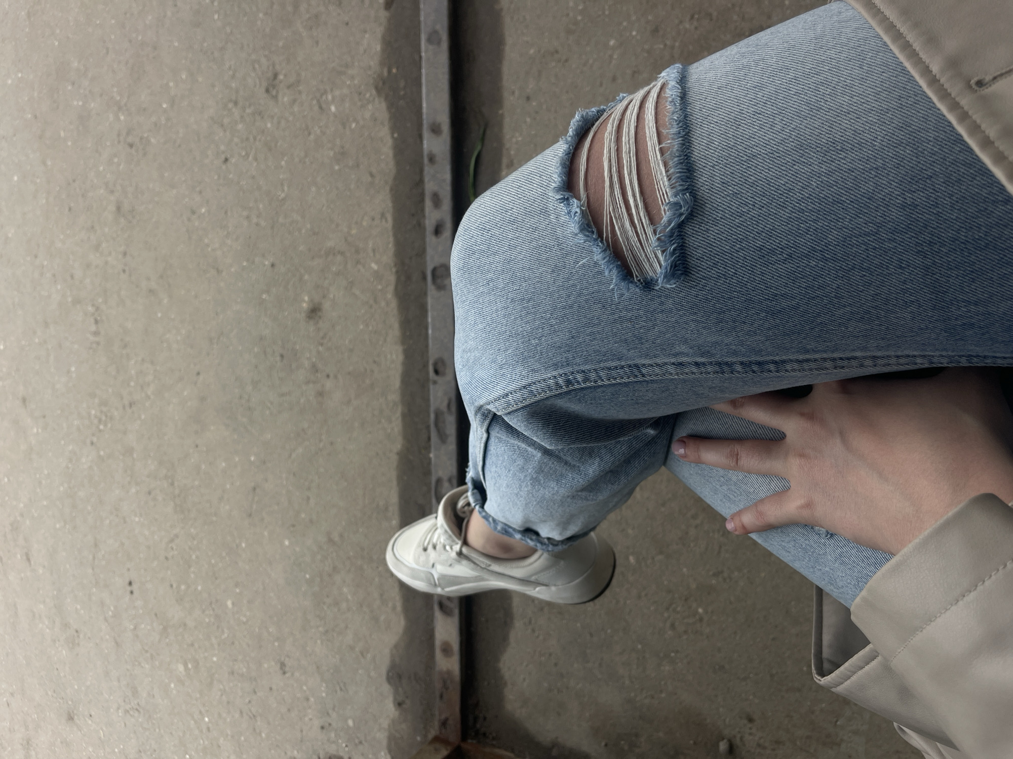 The image depicts a person seated on a concrete surface next to a corroded metal structure. The person is wearing light blue ripped jeans and white sneakers. Their right hand rests on their left leg. A beige jacket is visible. The location in Molokovo, Russia is not possible to verify from the image.