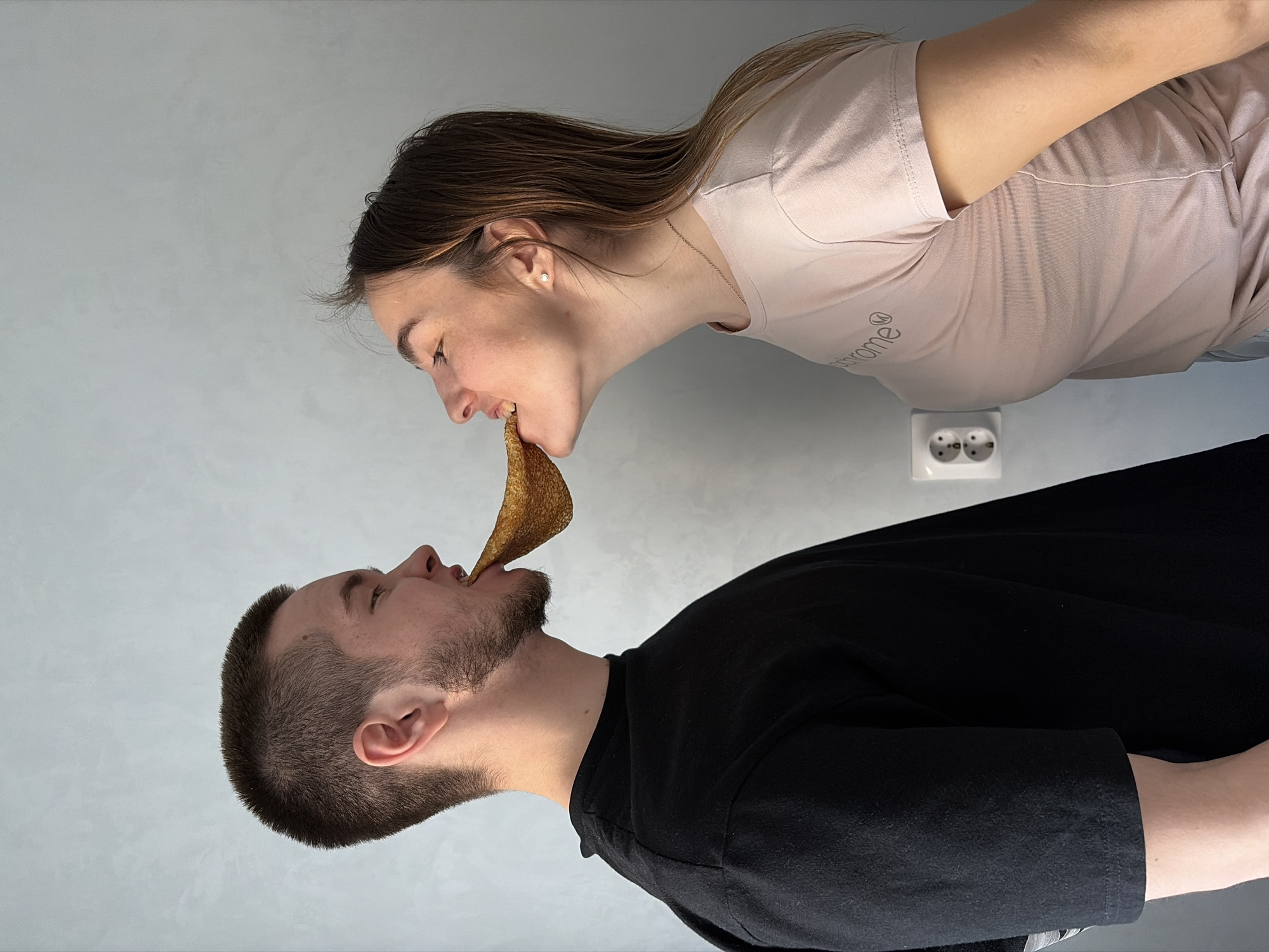 A man and a woman are sharing a pancake in what appears to be an indoor setting. The woman, wearing a light pink t-shirt, is positioned at the top of the image and is holding the upper part of the pancake with her mouth. The man, wearing a black t-shirt, is at the bottom of the image and is holding the lower part of the pancake with his mouth. A white electrical outlet is visible on the wall in the background, slightly below the woman. The background wall is light grey. The location is Unknown, Unknown.