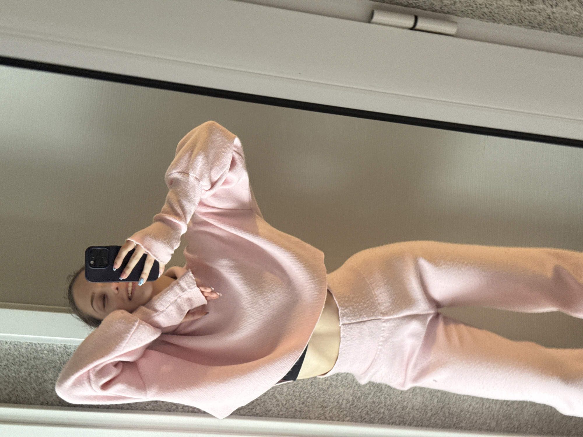 A young woman is taking a selfie in a mirror. She is wearing a light pink sweater and matching pants, and a black undergarment is visible at her waist. Her face is partially obscured by her phone, which she is holding with her left hand. Her nails are painted with blue and white designs.
The background appears to be a room with light-colored walls and carpet. The top of the mirror frame is visible at the top of the image, and what appear to be hinges can be seen above it.
The location is not discernible as Sofia, Bulgaria, based on the provided image. There are no distinctive landmarks or architectural features to identify the specific city.