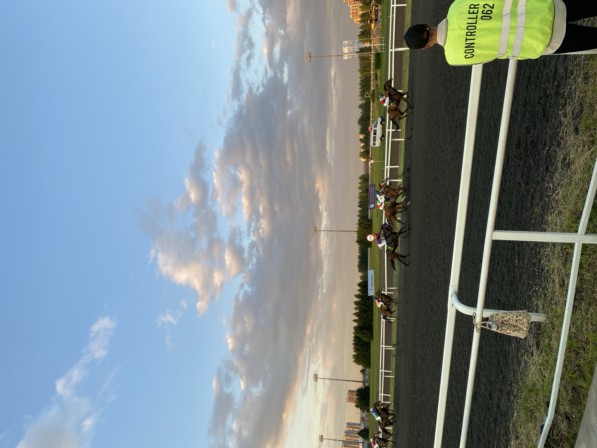 The image depicts a horse race at the Kazan Hippodrome in Kazan, Russia. The scene is captured during what appears to be late afternoon or early evening, with a sky filled with dramatic clouds tinged with pink and orange from the setting sun. Several horses and jockeys are actively racing on the dark, dirt track. In the foreground, a person wearing a bright yellow vest with "CONTROLLER 062" printed on the back is standing, facing away from the camera and towards the racetrack. A patterned bag is hanging from a white railing in the middle ground. The background shows a treeline, stadium lights, and buildings, suggesting an urban or semi-urban setting typical of a racecourse.
