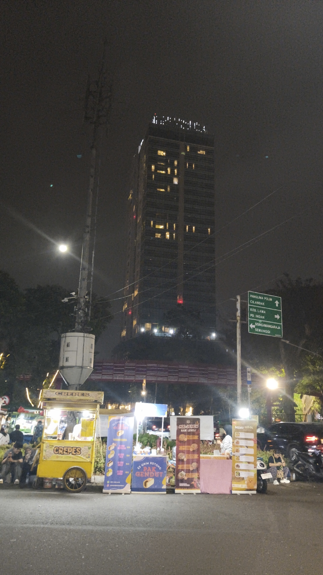 The image depicts a nighttime street scene in the city of South Jakarta, Indonesia. Dominating the background is a tall, modern skyscraper with illuminated windows, suggesting it is a hotel or office building. To the left of the skyscraper is a tall antenna tower. In the foreground, several food stalls are set up along the roadside. One prominent stall is a yellow crepes cart. Other stalls display various food items and menus, including what appears to be ice cream and noodles. People are visible around the stalls, some sitting and others standing. The street is paved, and there are trees and streetlights visible. A green directional signpost is also present, indicating directions to "PANGILMA POLIM CILANDAK," "KEB. LAMA," "PD. INDAH," and "SISINGAMANGARAJA SEMANGGI." The overall atmosphere suggests a bustling urban environment at night.