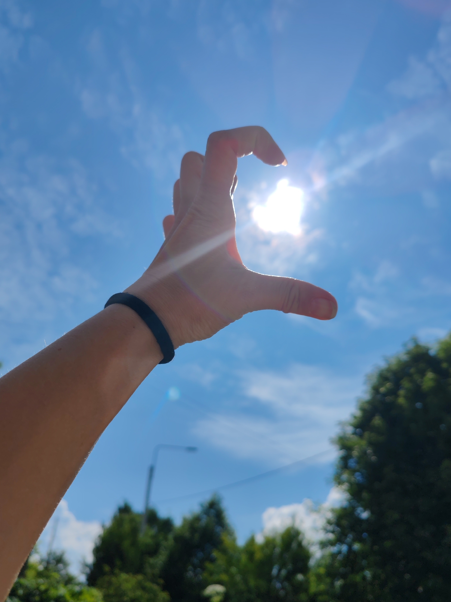 The image shows a close-up of a person's arm and hand against a bright blue sky with scattered clouds. The hand is held up, with the fingers curved in a way that suggests they are framing or interacting with the sun, which is visible and shining brightly, creating lens flares and streaks of light. A black wristband is worn on the arm. In the background, out of focus, are the tops of green trees and a streetlamp, indicating an outdoor setting. The bright sunlight suggests it is daytime, likely a clear or partly cloudy day. The overall impression is one of a sunny, possibly warm, day. There is no visible text in the image.