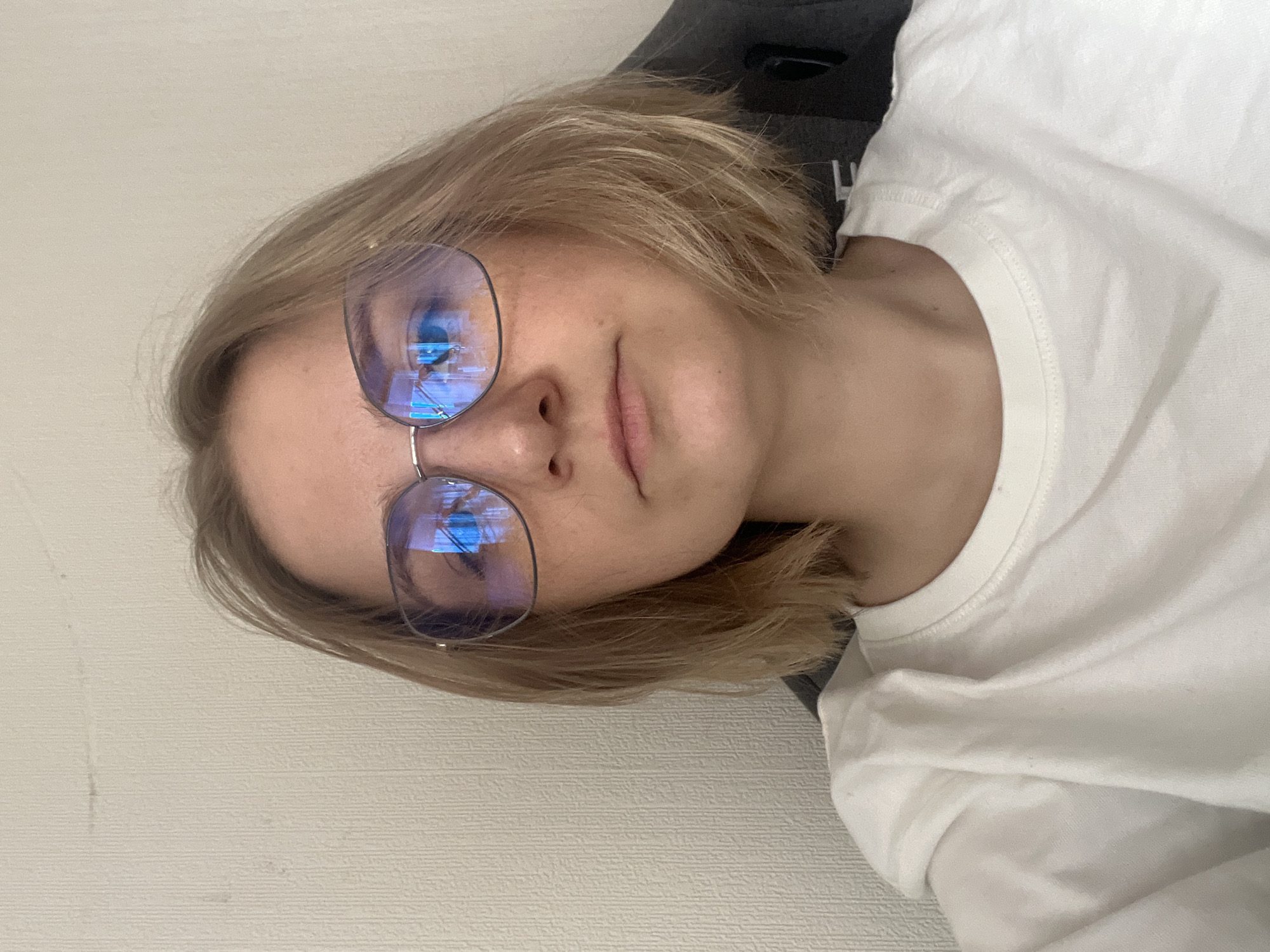 A fair-skinned individual with short, blonde hair and wearing eyeglasses is visible in this image. They are wearing a white T-shirt and appear to be seated in a chair, with a black headrest partially visible behind them. The setting appears to be indoors, with a light-colored wall in the background. The location in Novosibirsk, Russia, cannot be verified.