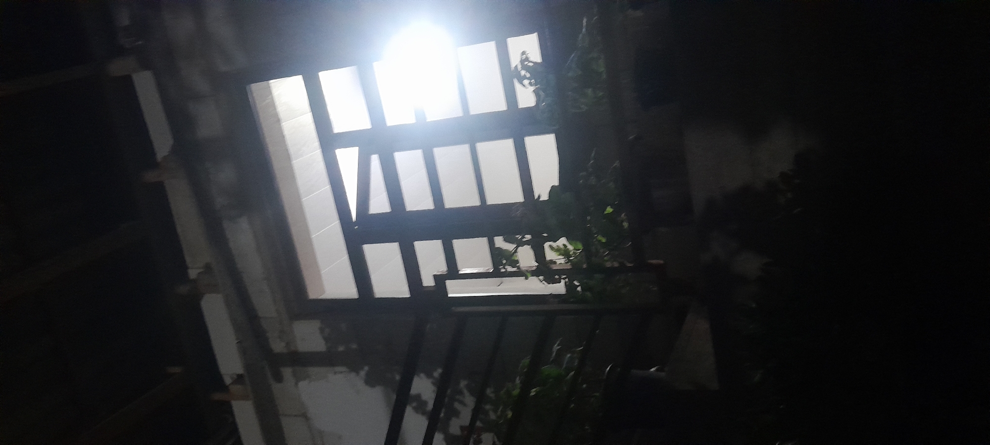 The image depicts a view from below of a building's exterior at night. A large window, divided into multiple rectangular panes by dark framing, is visible. The window is illuminated from within by a bright light source, creating a glare. Several dark, leafy plants in pots are situated on a ledge or balcony in front of the window, casting shadows. The building appears to be constructed of concrete blocks. The scene is dimly lit, with the foreground and the area above the window almost completely obscured by darkness. Wooden beams are visible extending from a dark ceiling structure above the window. The context of Chartak, Uzbekistan, suggests this might be an outdoor area of a residential or commercial building in that location.