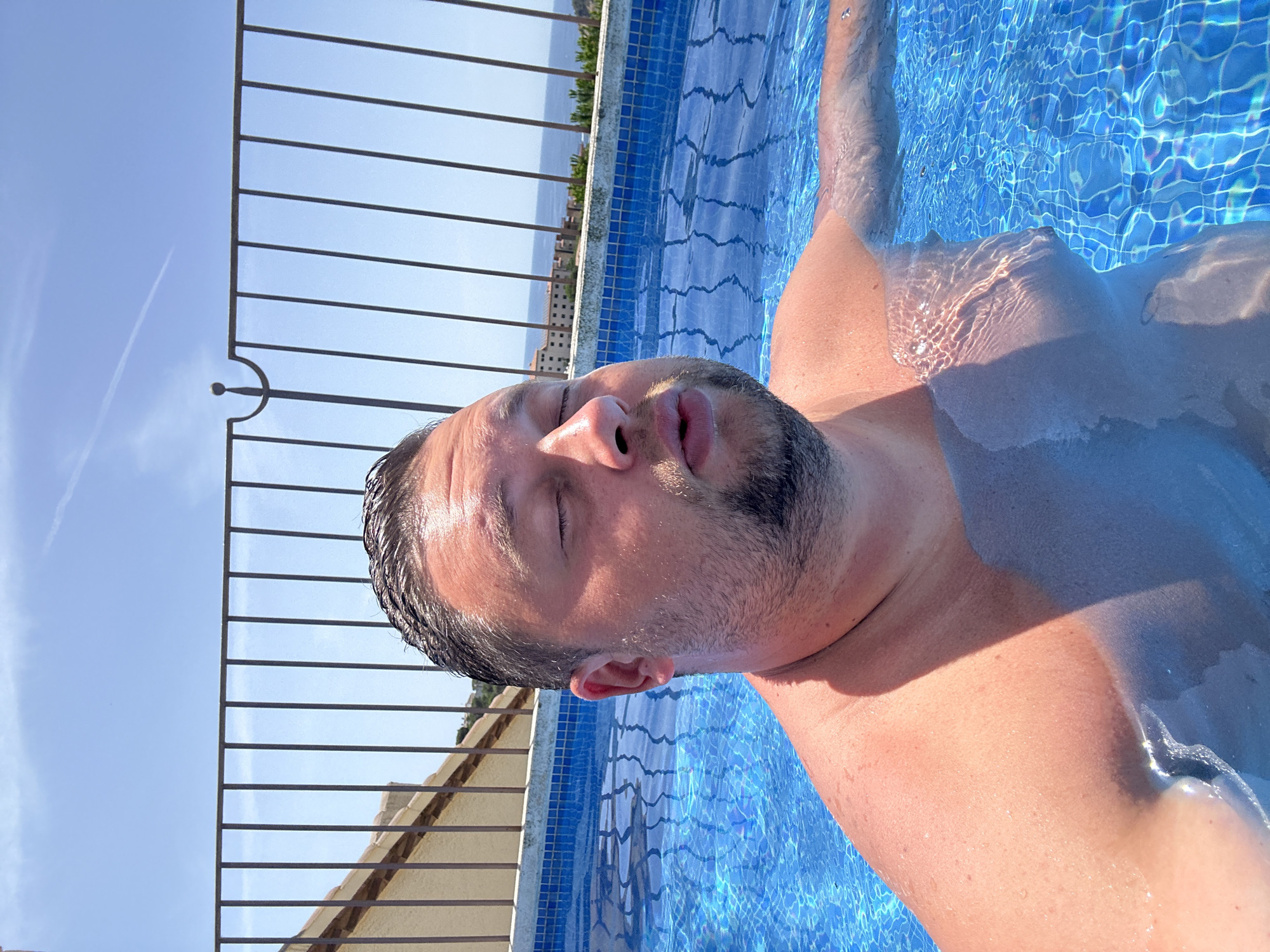 A man is in a swimming pool on a sunny day. He has his eyes closed and his mouth slightly open. The pool is surrounded by a tiled edge and a metal railing. In the background, there are buildings and the sea. The image appears to be taken from a high vantage point, possibly a rooftop pool in Sant Feliu de Guíxols, Spain, given the architectural style and coastal view.